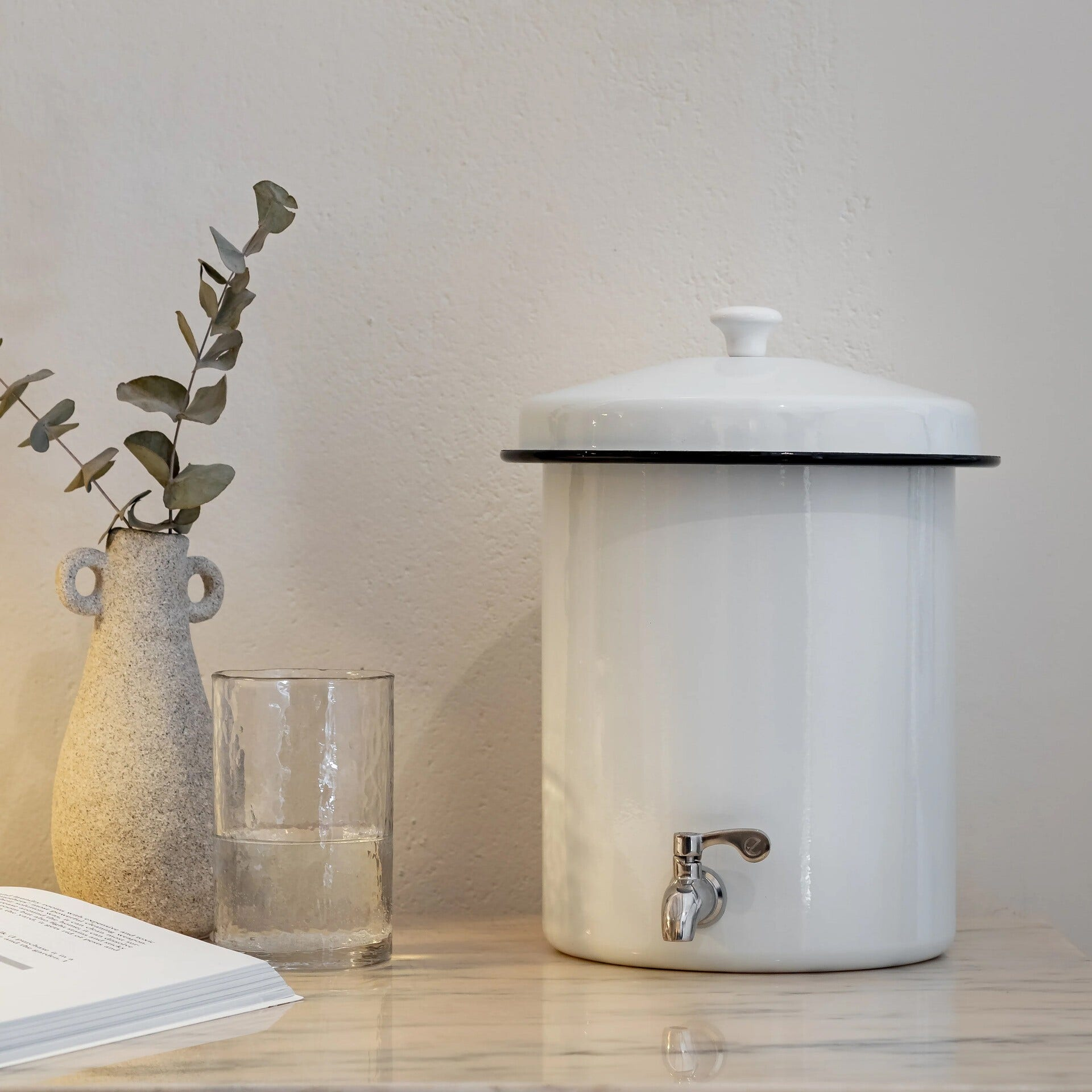 Distributore d'acqua bianco con bicchiere, vaso con piante e libro su superficie in marmo.