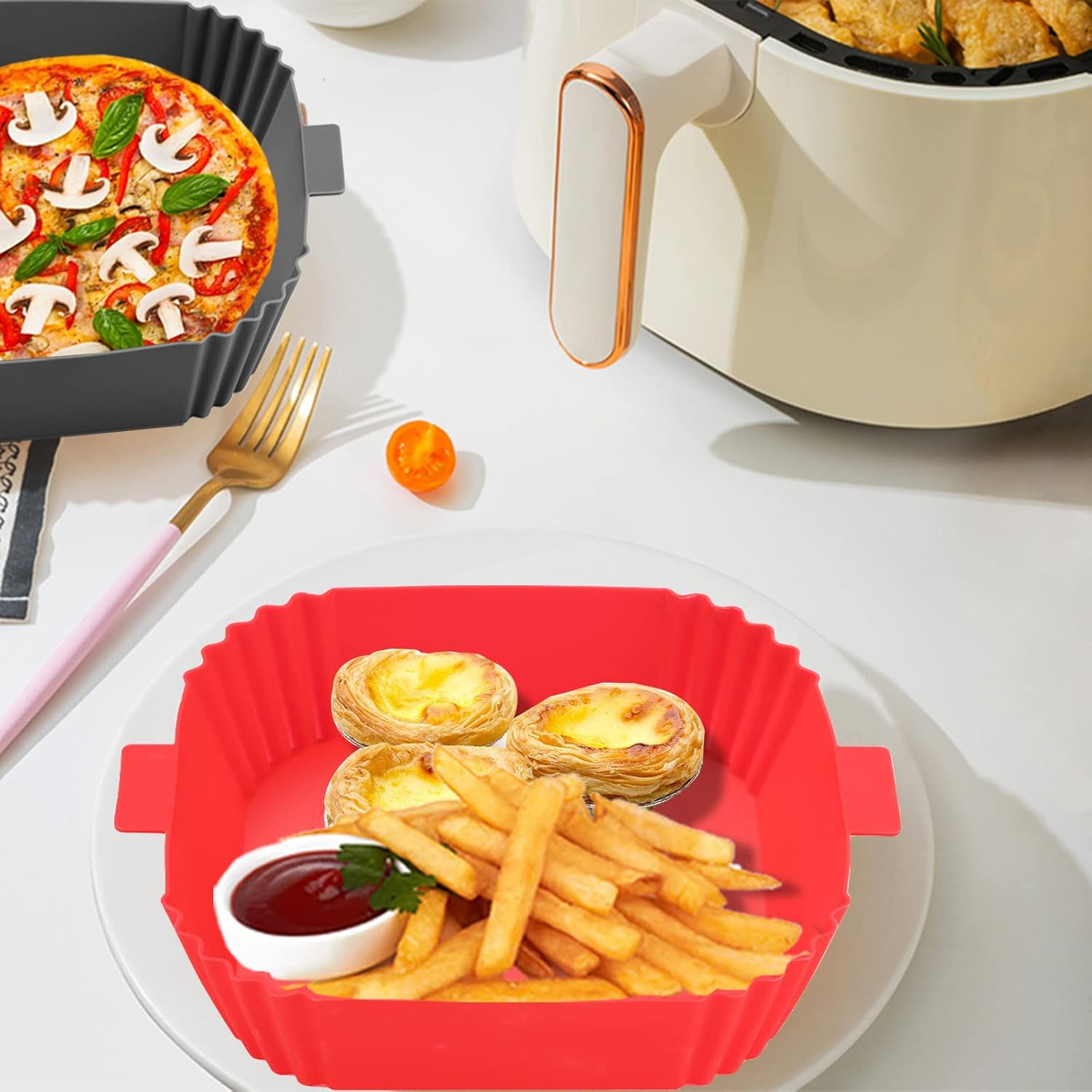 Ein rotes Silikonfach mit Pommes, Eiertörtchen und Ketchup auf einem weißen Teller. Eine Pizza in einem schwarzen Fach und eine Heißluftfritteuse sind sichtbar.
