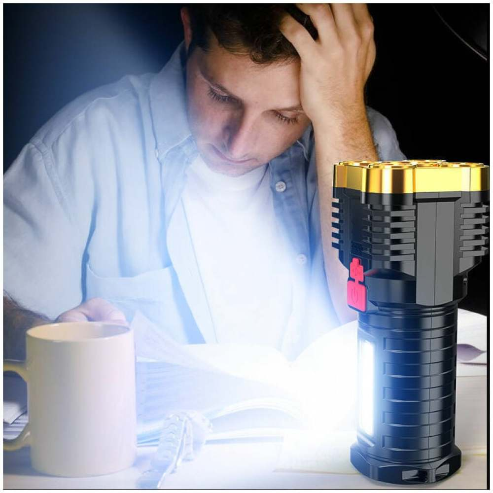 Mann mit Hand am Kopf, liest bei Taschenlampenlicht. Taschenlampe ist schwarz und gold. Becher und Papiere auf dem Tisch. Szene ist dunkel.