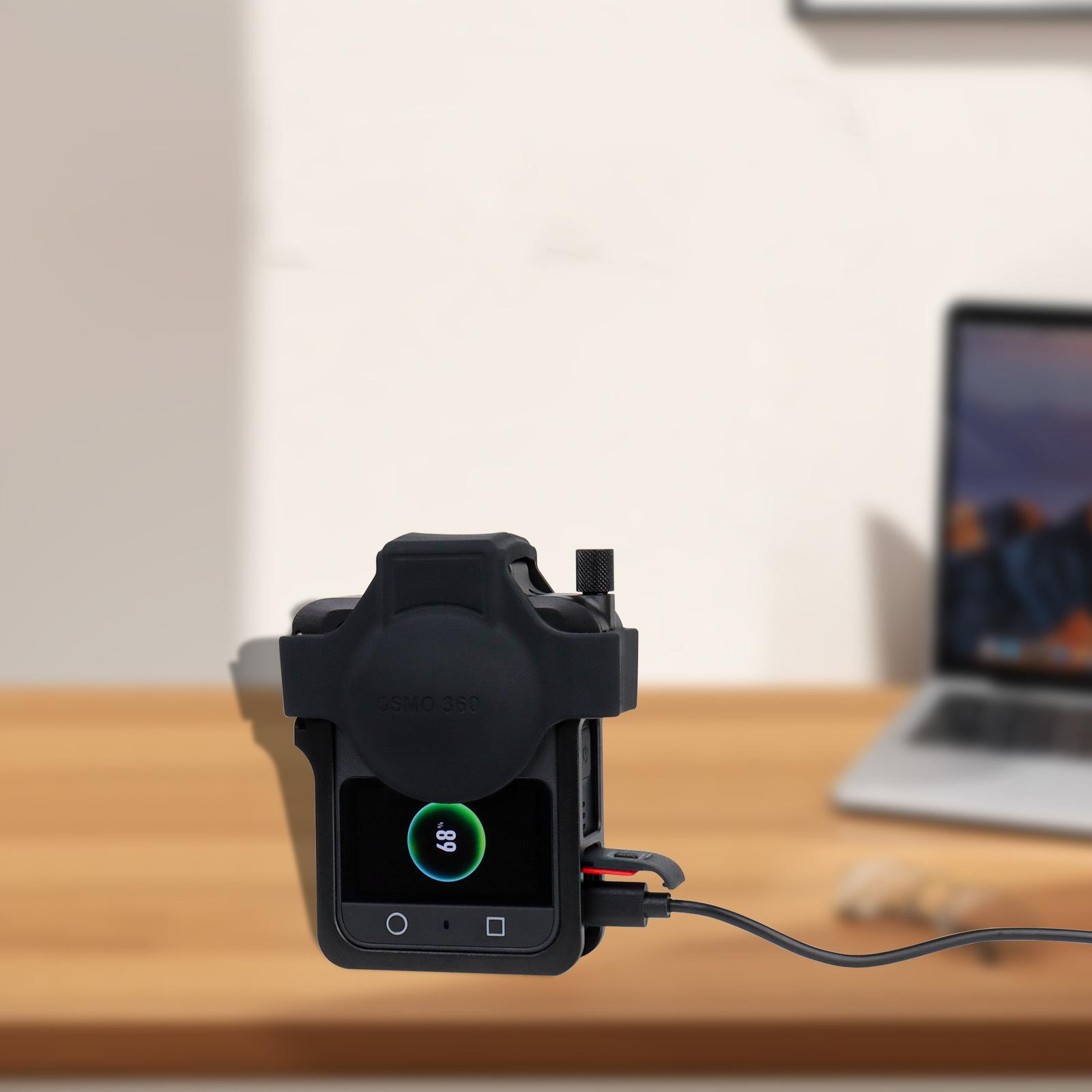 Eine schwarze Kamera wird aufgeladen und zeigt 68 % Akku an. Sie steht auf einem Holztisch, im Hintergrund ein Laptop.