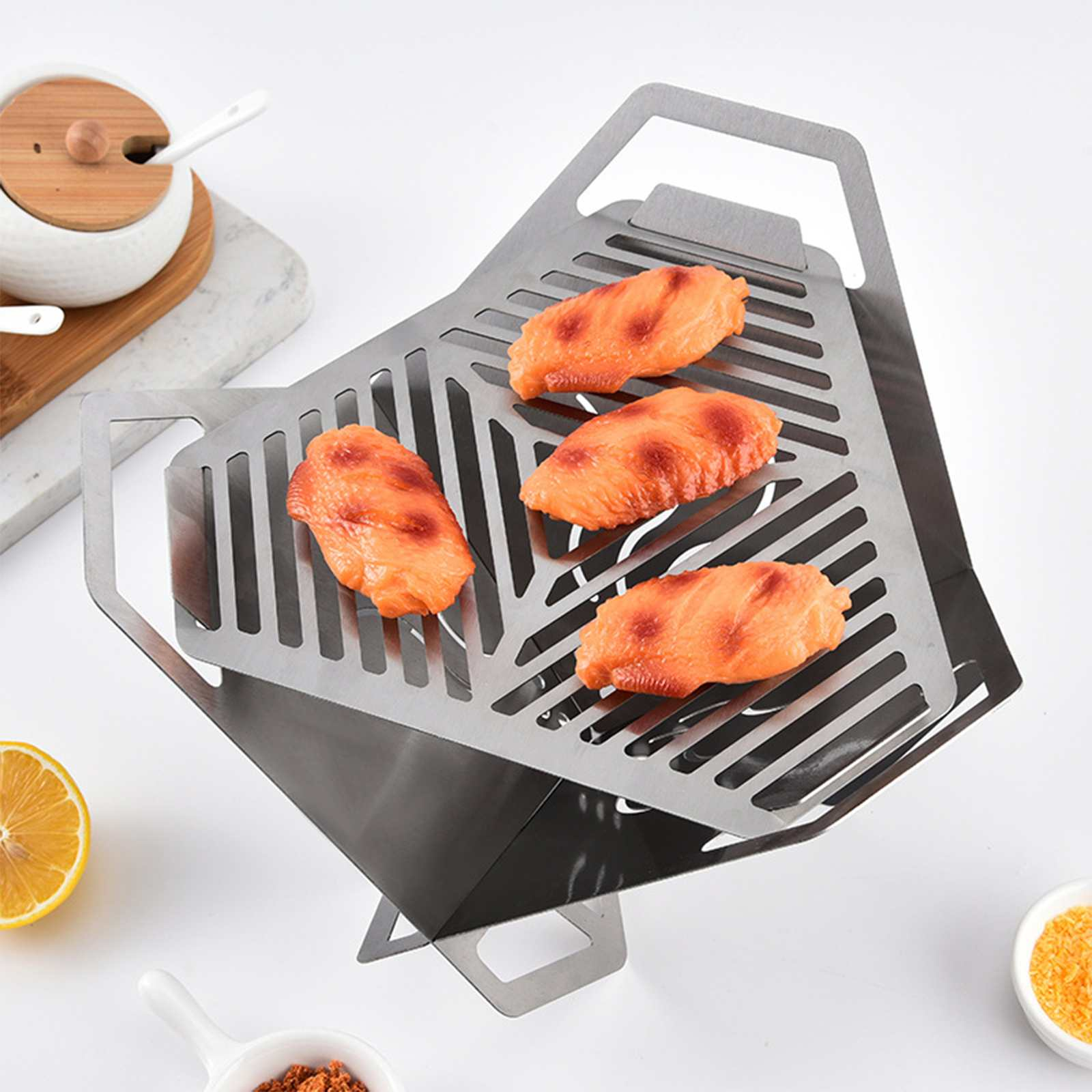 Ein Metallgrill mit Hähnchenflügeln. Enthält eine Zitrone, Gewürze und eine kleine Schüssel mit Deckel. Alles auf weißem Hintergrund.