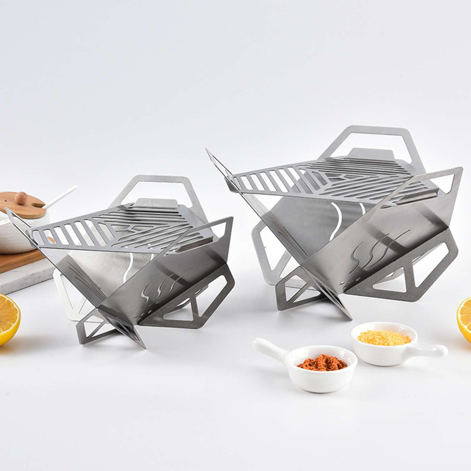 Zwei tragbare Metallgrills. Schalen mit Gewürzen und Zitronenhälften im Vordergrund. Weißer Hintergrund.