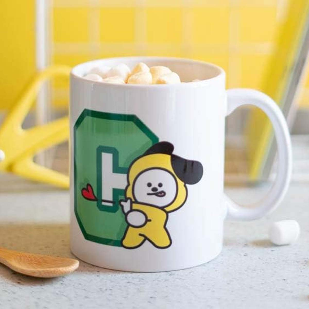 Eine weiße Tasse mit einem Cartoon-Charakter, Chimmy, gefüllt mit Marshmallows. Gelbe und grüne Akzente mit einem Holzlöffel und einem Marshmallow.