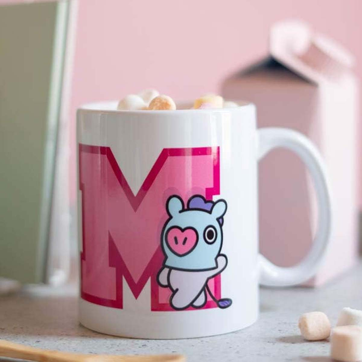 Eine weiße Tasse mit einem rosa 'M' und einer Cartoon-Figur. Marshmallows darin, mit einer rosa Box und einem Buch im Hintergrund.
