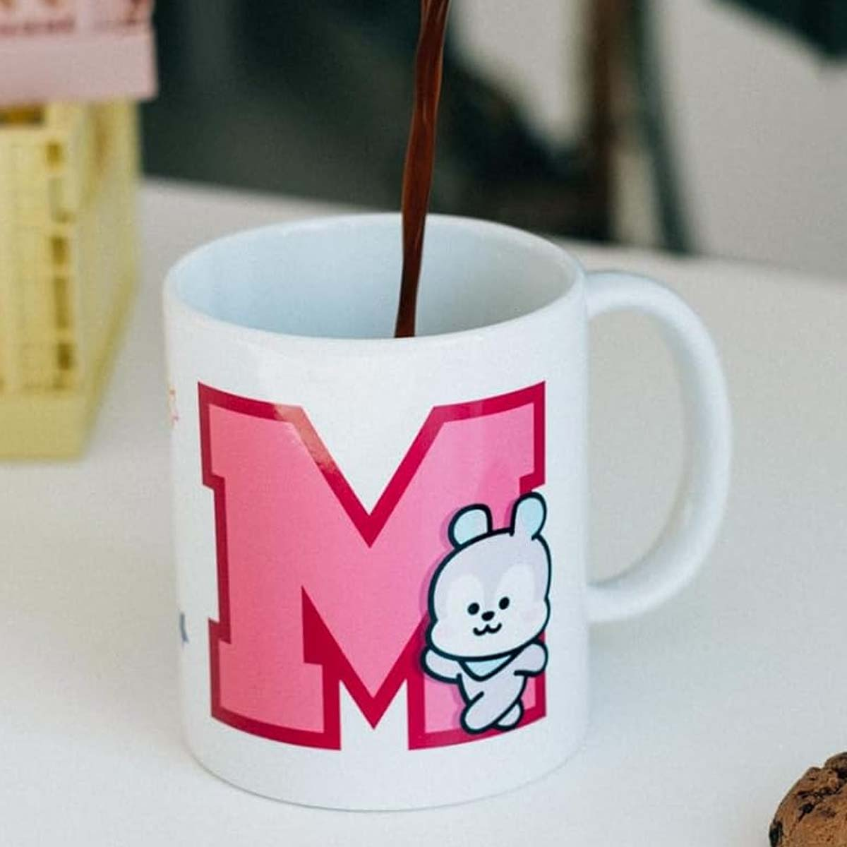 Eine weiße Tasse mit rosa Buchstaben 'M' und einem Cartoon-Bären wird mit brauner Flüssigkeit gefüllt.
