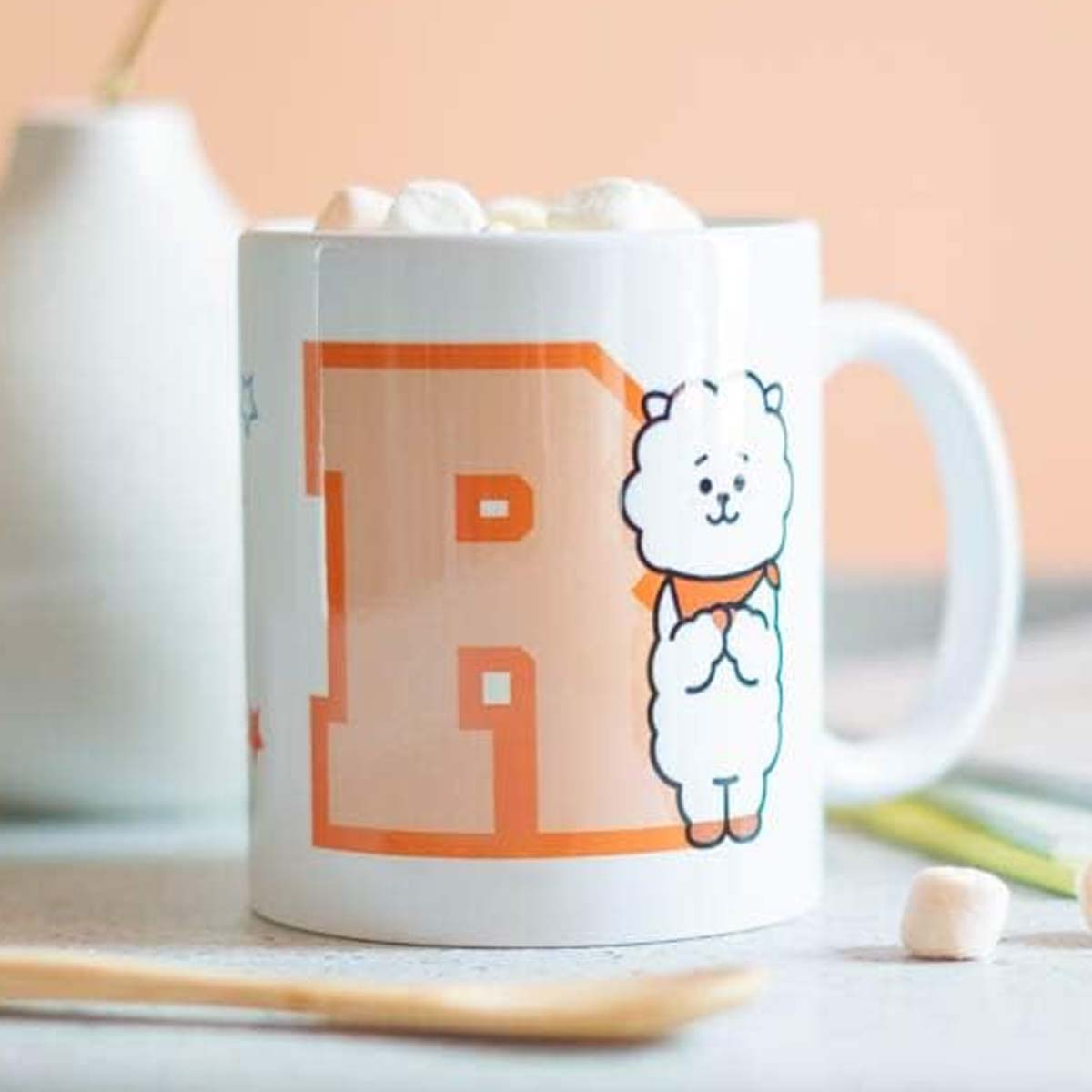 Weißer Becher mit orangefarbenem 'R' und Cartoon-Figur, gefüllt mit Marshmallows. Holzlöffel und Vase im Hintergrund.