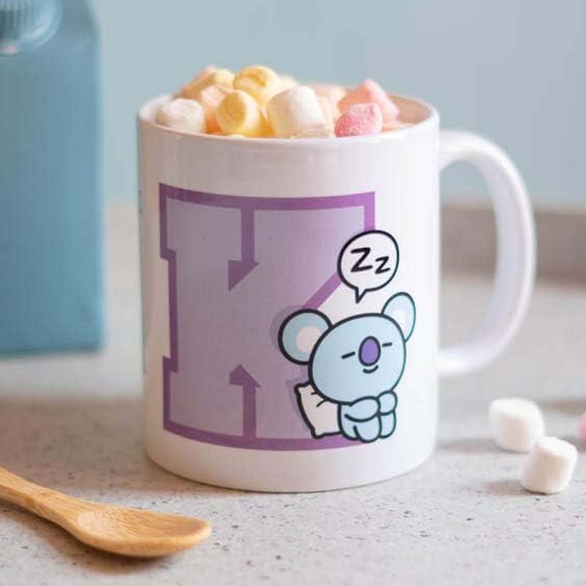 Eine weiße Tasse mit einem Koala-Cartoon und Marshmallows. Holzlöffel links. Blauer Behälter im Hintergrund.