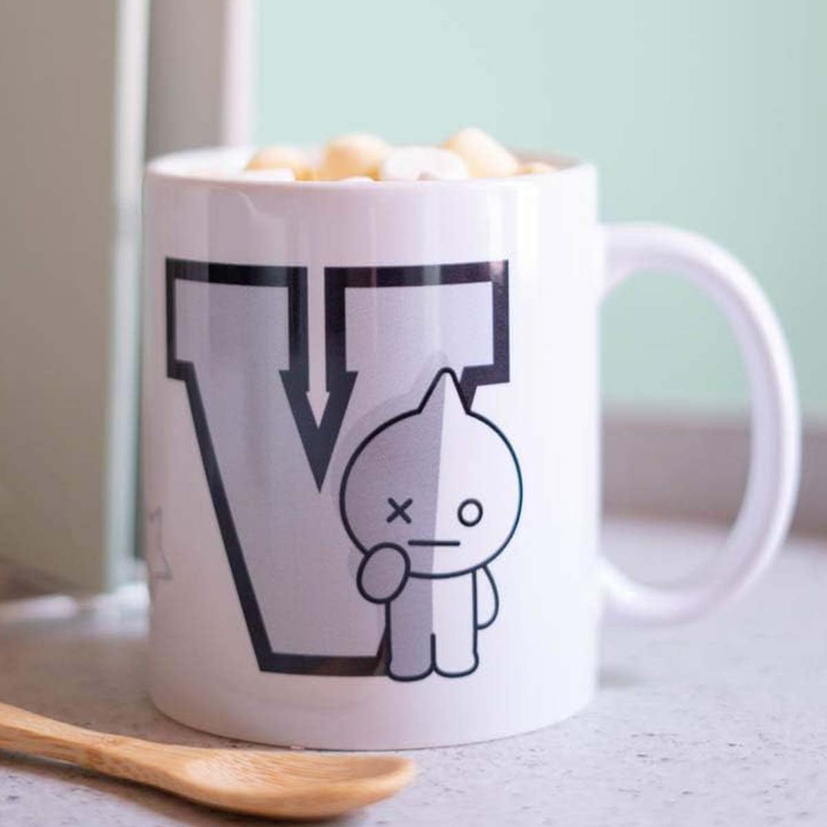 Eine weiße Tasse mit einem Cartoon-Charakter und dem Buchstaben V, gefüllt mit Marshmallows, steht neben einem Holzlöffel.