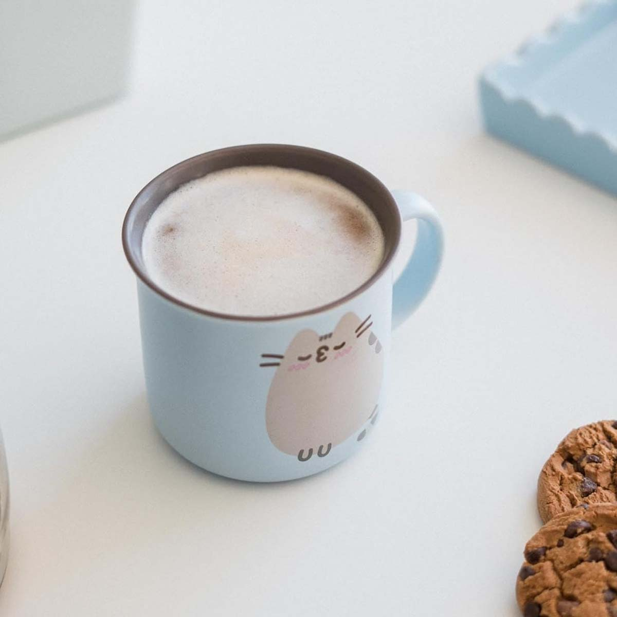 Hellblauer Becher mit Katzenillustration, gefüllt mit schaumigem Getränk, steht auf weißer Oberfläche mit Keksen.