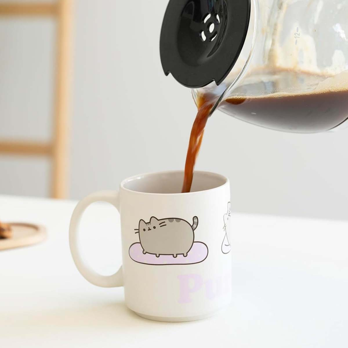 Kaffee wird in eine weiße Tasse mit Katzendesign gegossen. Die Tasse steht auf einem Tisch. Der Kaffeezubereiter befindet sich über der Tasse.