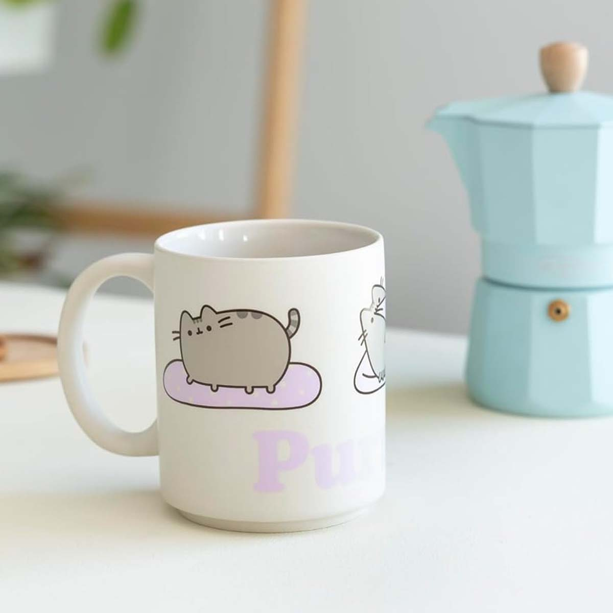 Eine weiße Tasse mit einer Katzenillustration und dem Wort 'Pusheen' daneben, neben einem blauen Kaffeezubereiter auf weißer Oberfläche.