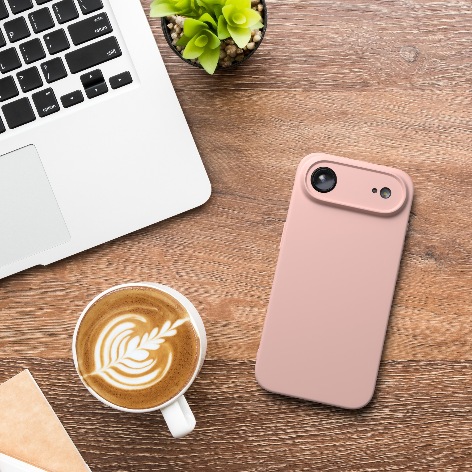 Auf Holz liegen ein Laptop, ein Telefon, Kaffee und eine Pflanze. Das Telefon ist rosa. Der Kaffee hat Latte Art.