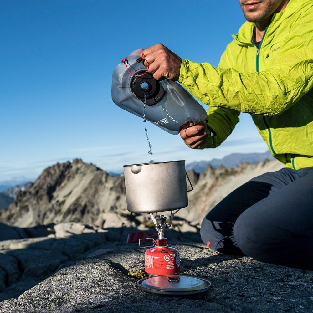 Eine Person gießt Wasser aus einem Trinkrucksack in einen Topf auf einem Campingkocher, der auf einem Felsen steht, mit Bergen im Hintergrund.