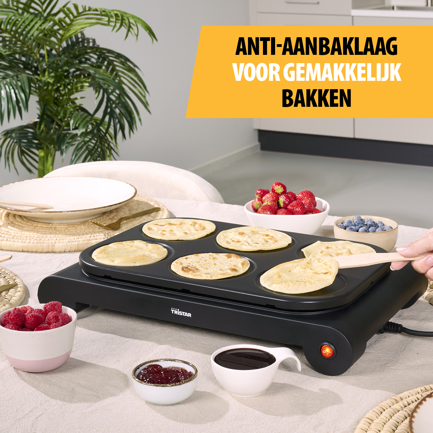 Ein Pfannkuchengrill mit Pfannkuchen, Beeren und Saucen auf einem Tisch. Eine Hand dreht einen Pfannkuchen mit einem Spatel.