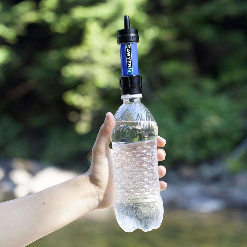 Eine Hand hält eine klare Wasserflasche mit einem blau-schwarzen Wasserfilter. Der Hintergrund zeigt verschwommene grüne Bäume und Wasser.