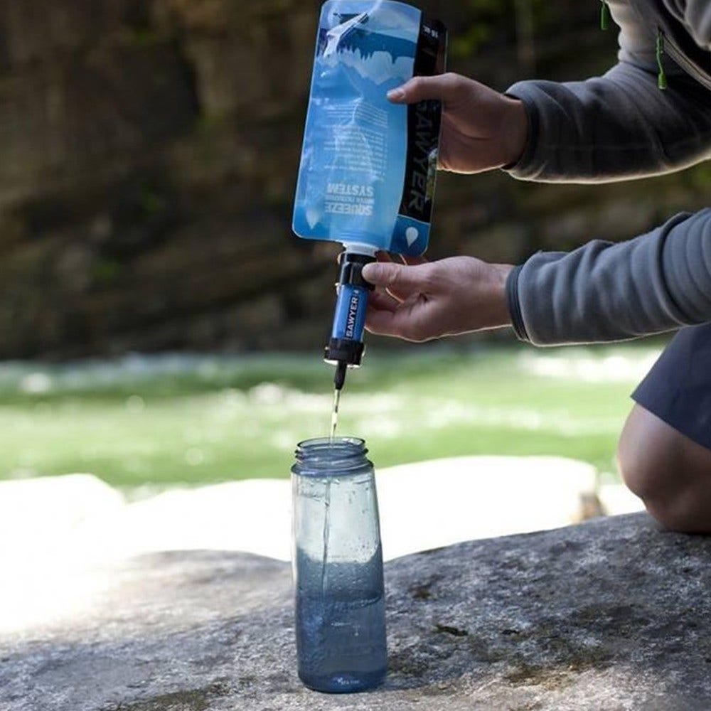 Eine Person filtert Wasser aus einem blauen Beutel in eine klare Flasche mit einem Sawyer-Filter, in der Nähe eines fließenden Flusses. Das Wasser ist klar.