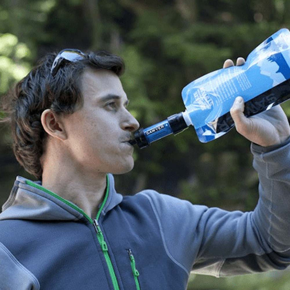 Ein Mann trinkt Wasser aus einer blauen Filterflasche. Er trägt eine graue Jacke und eine Sonnenbrille. Im Hintergrund sind grüne Bäume zu sehen.
