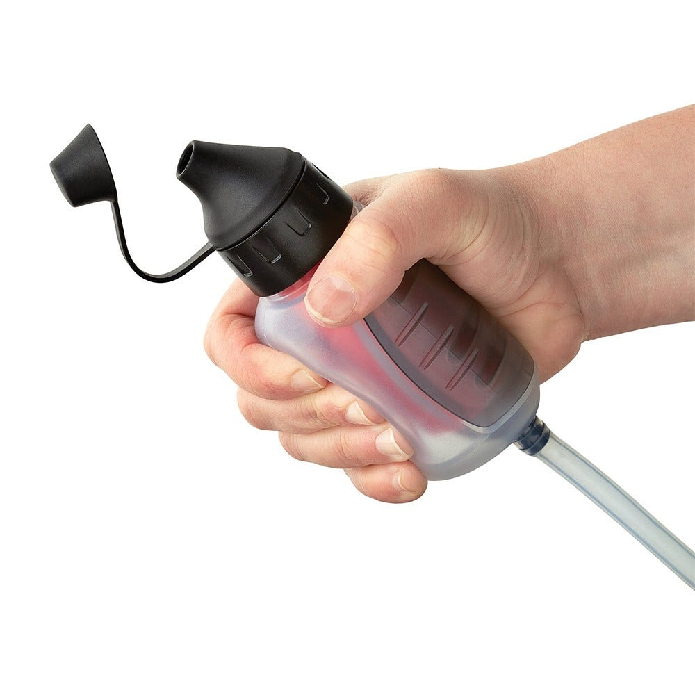 Hand hält eine Wasserflasche mit schwarzem Deckel. Ein klarer Schlauch erstreckt sich von der Flasche. Die Flasche ist teilweise mit roter Flüssigkeit gefüllt.