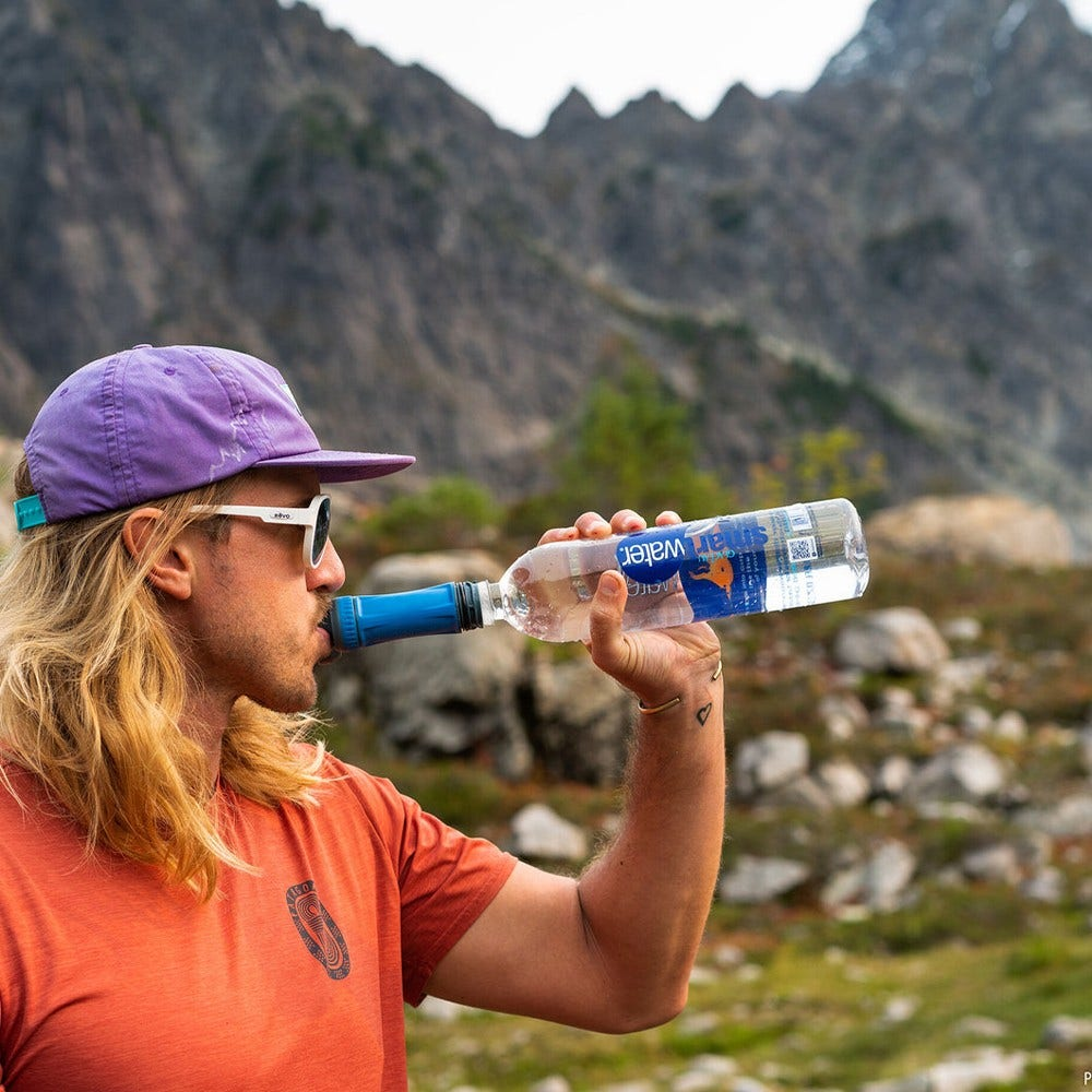 Ein Mann trinkt Wasser aus einer Smartwater-Flasche. Er trägt eine lila Kappe, Sonnenbrille und ein orangefarbenes Hemd. Berge sind im Hintergrund.