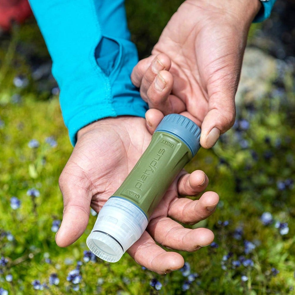 Hände halten einen grünen Platypus-Wasserfilter. Der Hintergrund zeigt grünes Gras und kleine lila Blumen. Die Person trägt eine blaue Jacke.
