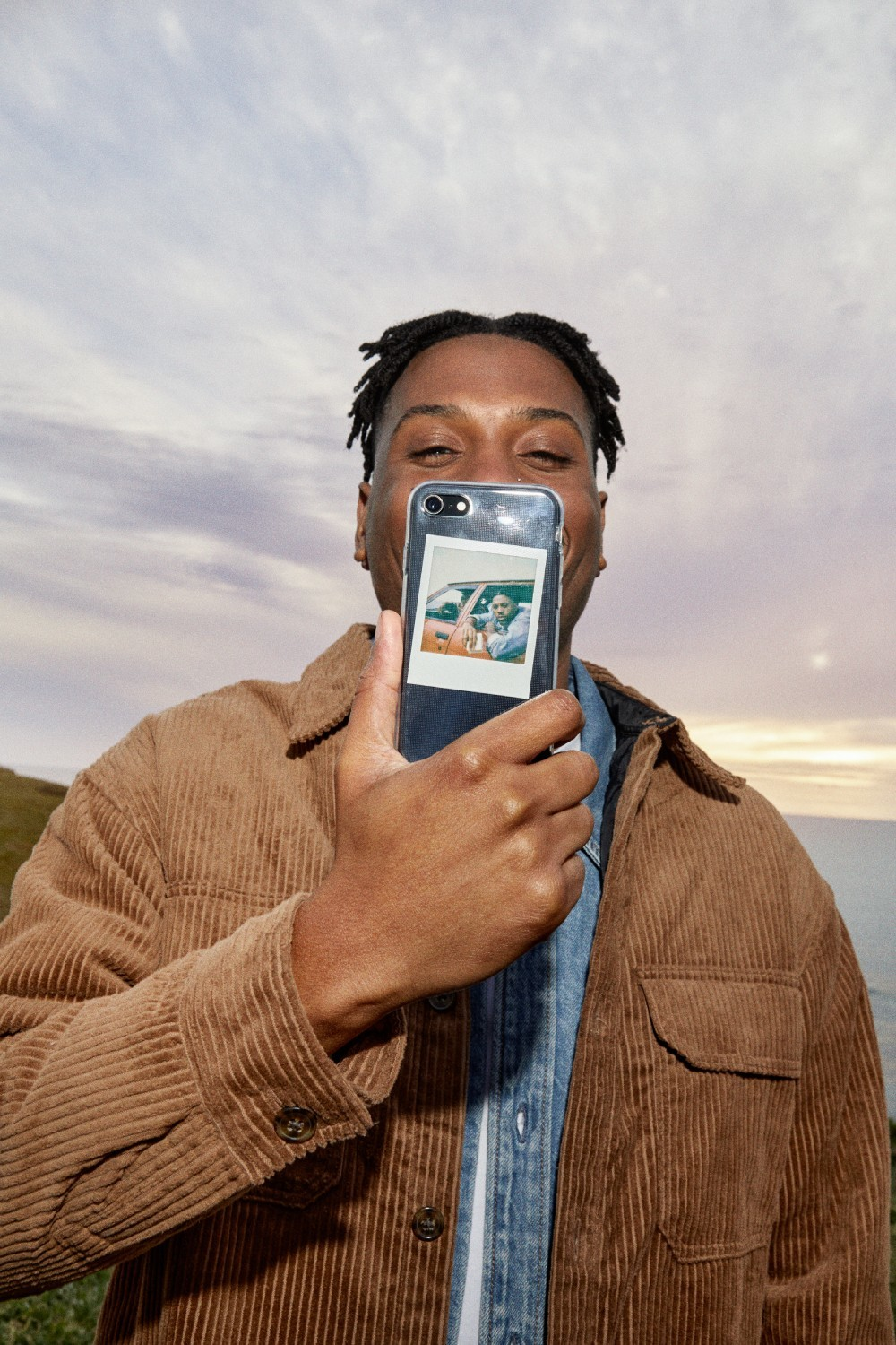 Ein Mann hält ein Telefon hoch, auf dem ein Foto zu sehen ist. Er trägt eine braune Cordjacke. Der Hintergrund zeigt einen bewölkten Himmel und Meer.