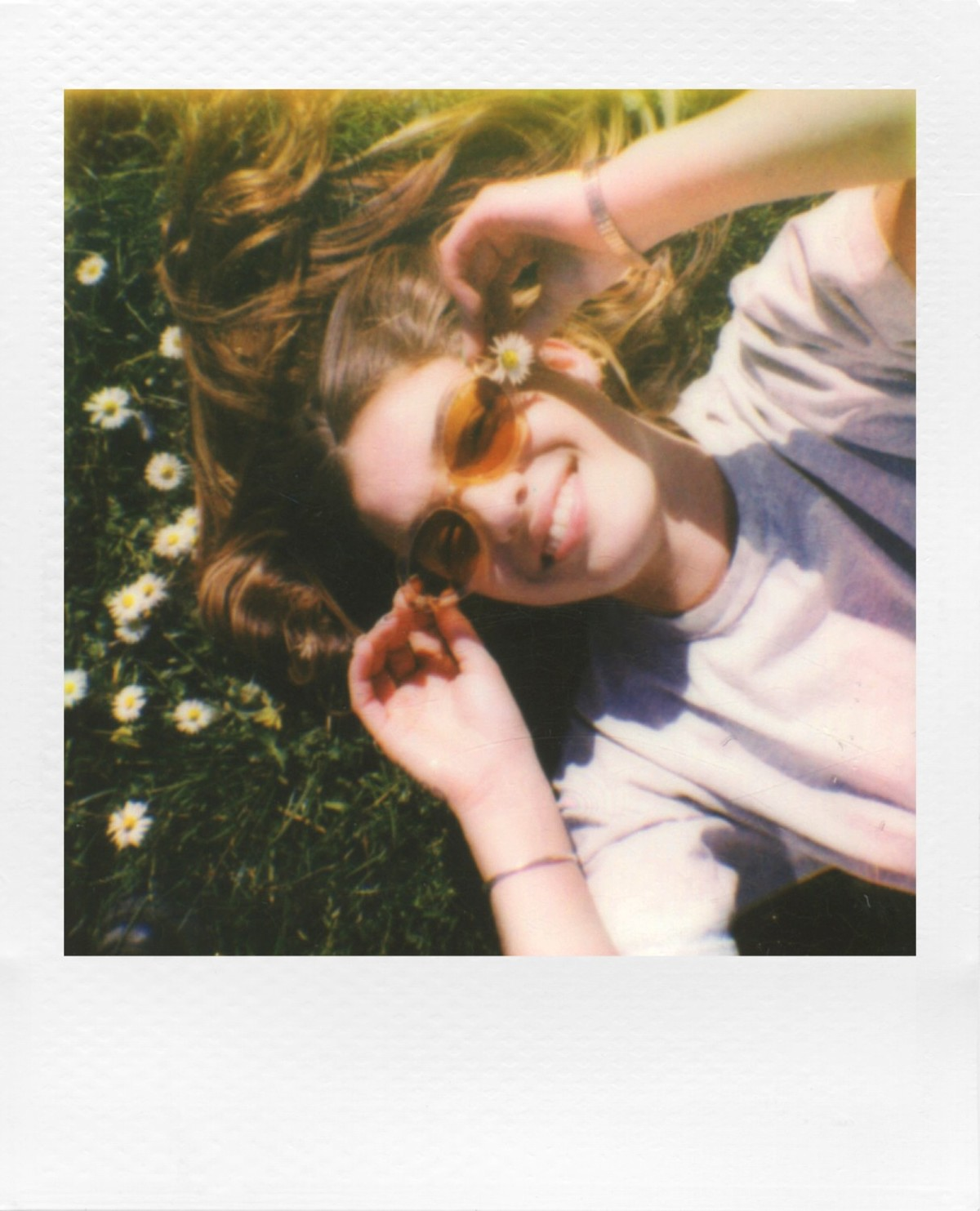 Eine lächelnde Frau liegt im Gras, trägt Sonnenbrille und hält Gänseblümchen. Sie ist von Blumen umgeben und genießt das Sonnenlicht.