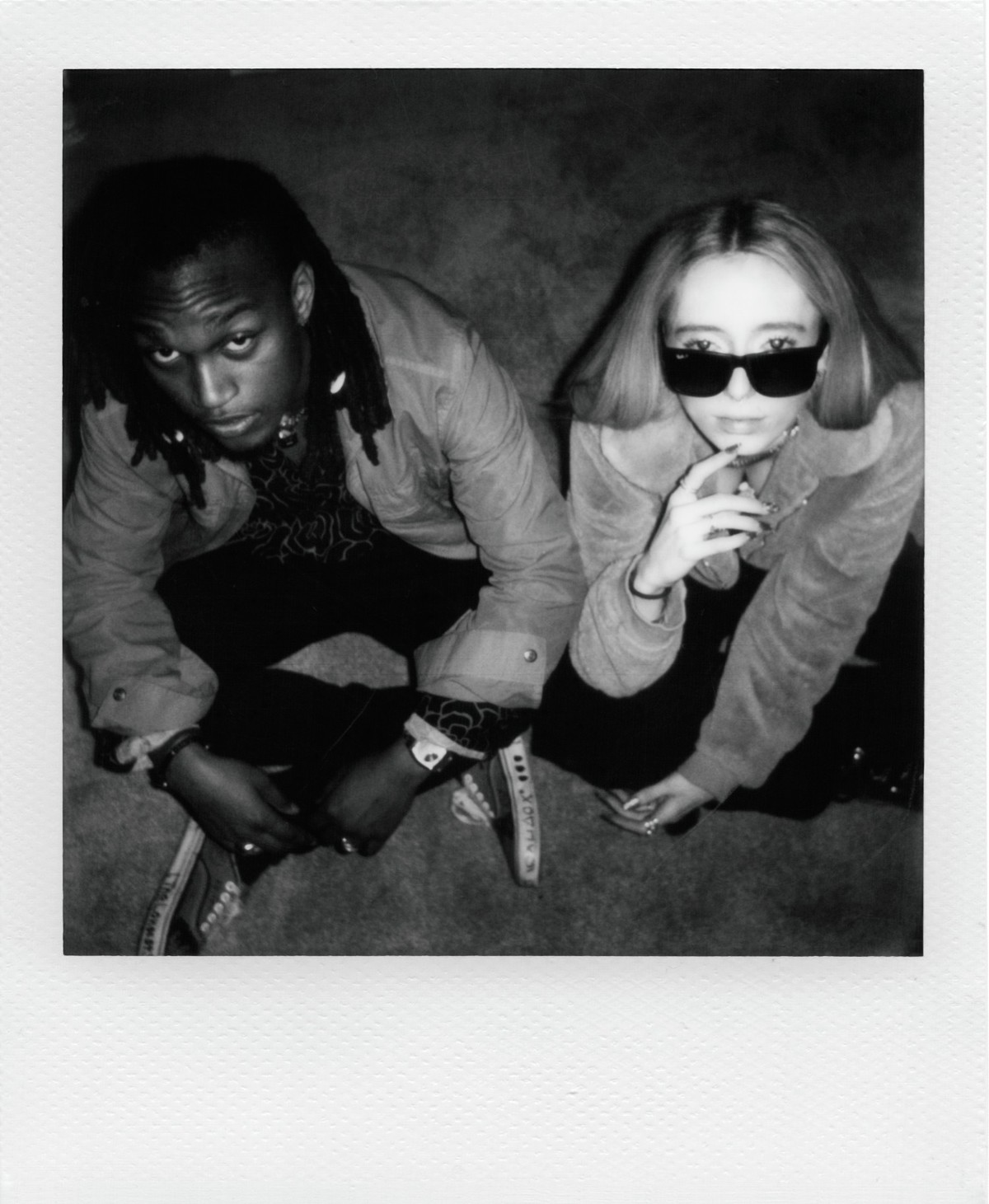 Czarno-białe zdjęcie dwóch osób, jedna z dredami, druga z okularami przeciwsłonecznymi, siedzących i patrzących w górę.