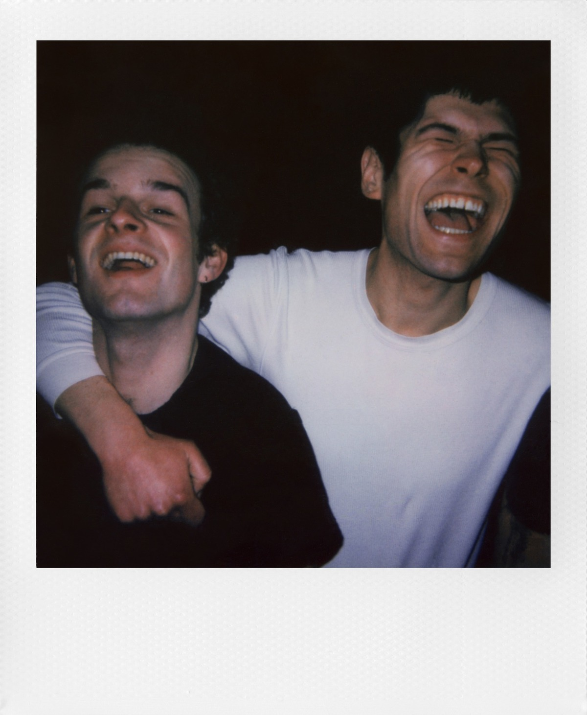 Twee mannen lachen, een met een arm om de ander. Eén draagt een wit shirt, de ander een zwart shirt. Donkere achtergrond.
