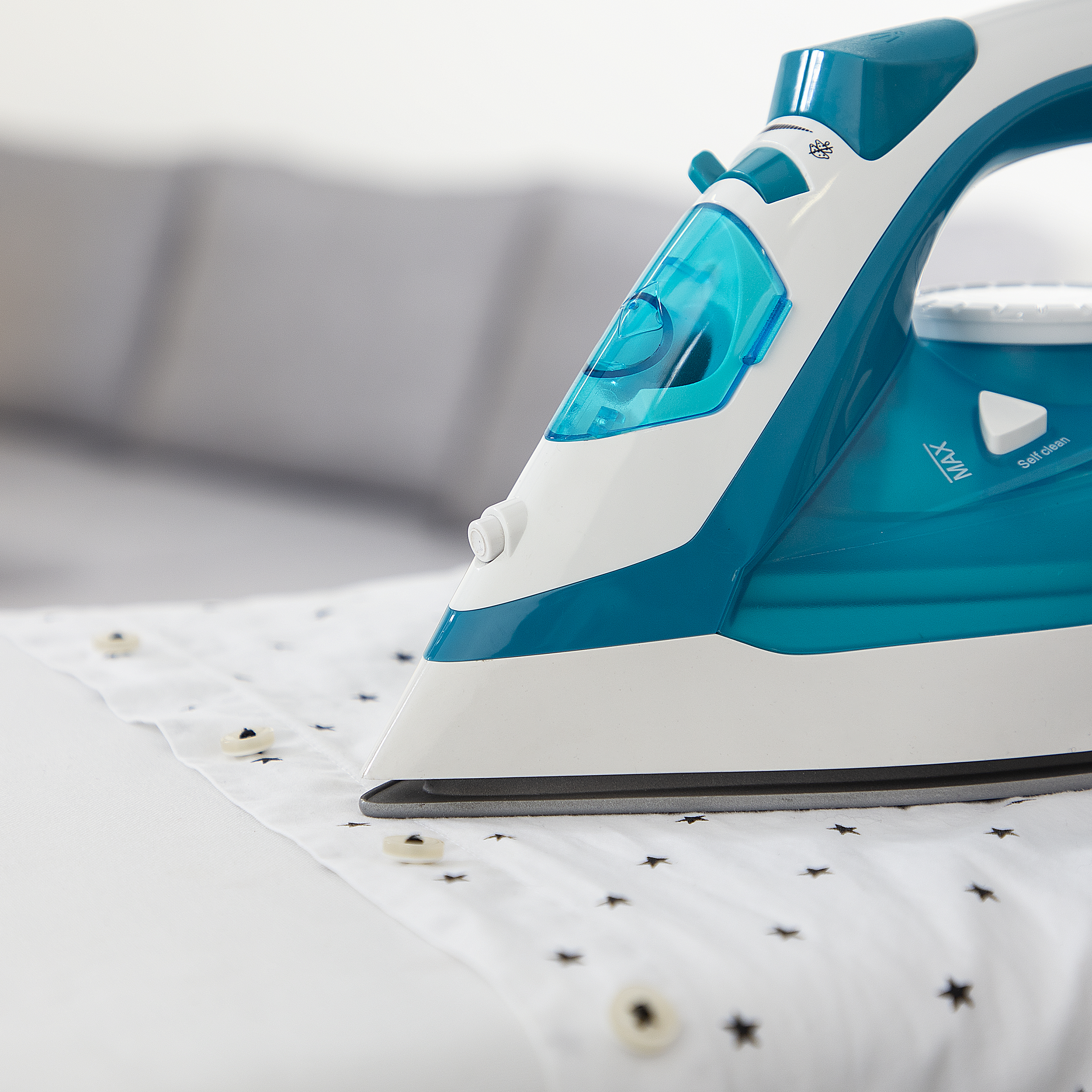 Niebiesko-białe żelazko na białej koszuli z czarnymi nadrukami gwiazd. Widoczne guziki. Tło jest rozmyte.