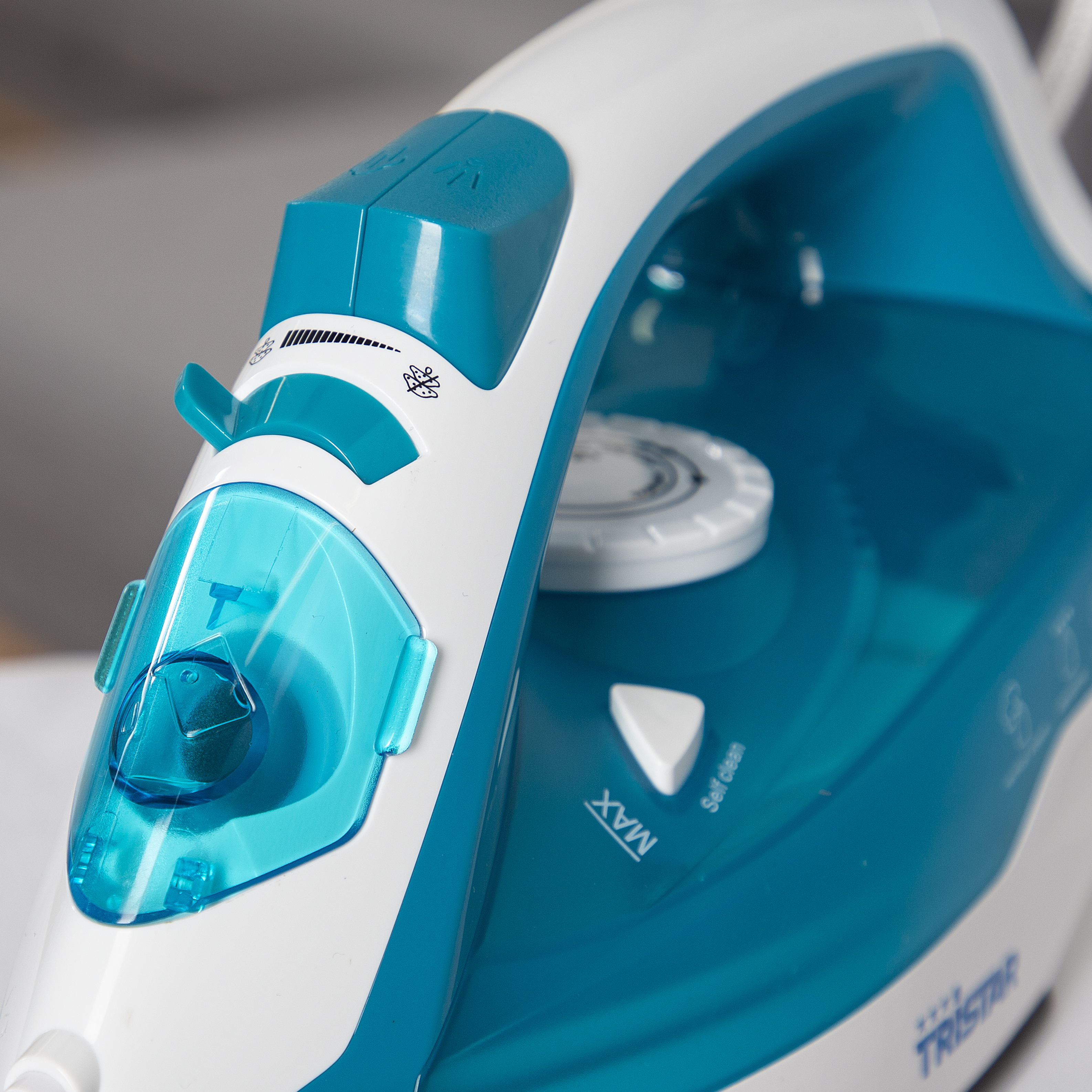 Zbliżenie białego i turkusowego żelazka. Widoczne są elementy sterujące i szczegóły marki. Żelazko leży na białej powierzchni.