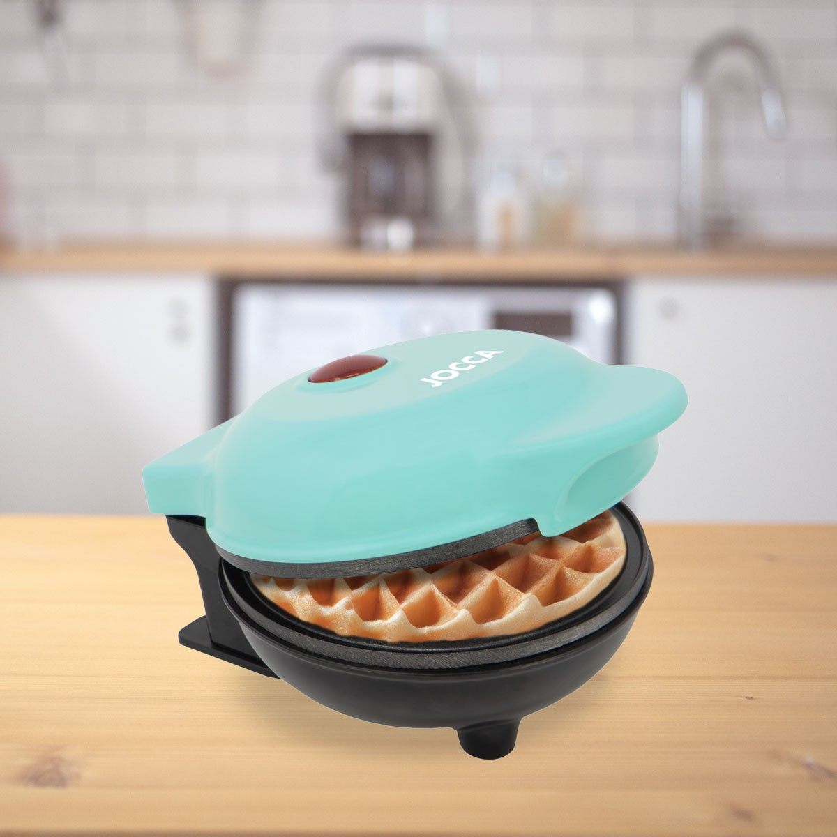 Una macchina per waffle turchese con un waffle all'interno è su un tavolo di legno. Lo sfondo mostra una cucina con elettrodomestici.
