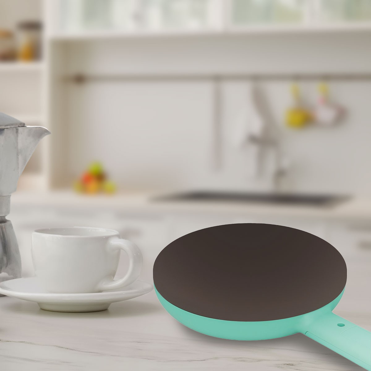 Una escena de cocina con una taza de café, una crepera y una cafetera. El fondo está ligeramente borroso.