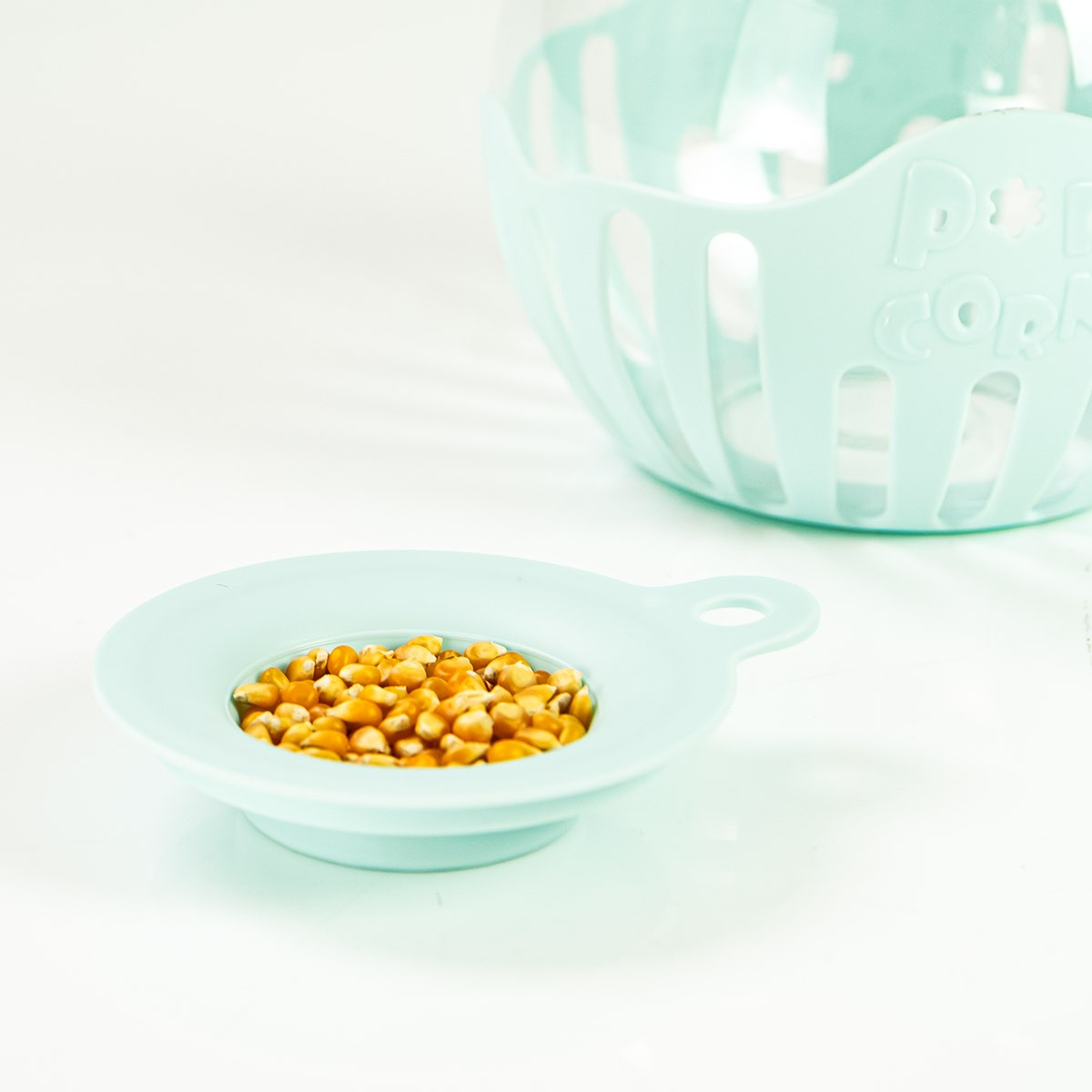 Una piccola ciotola blu con chicchi di mais. Sullo sfondo, una macchina per popcorn blu più grande su una superficie bianca.