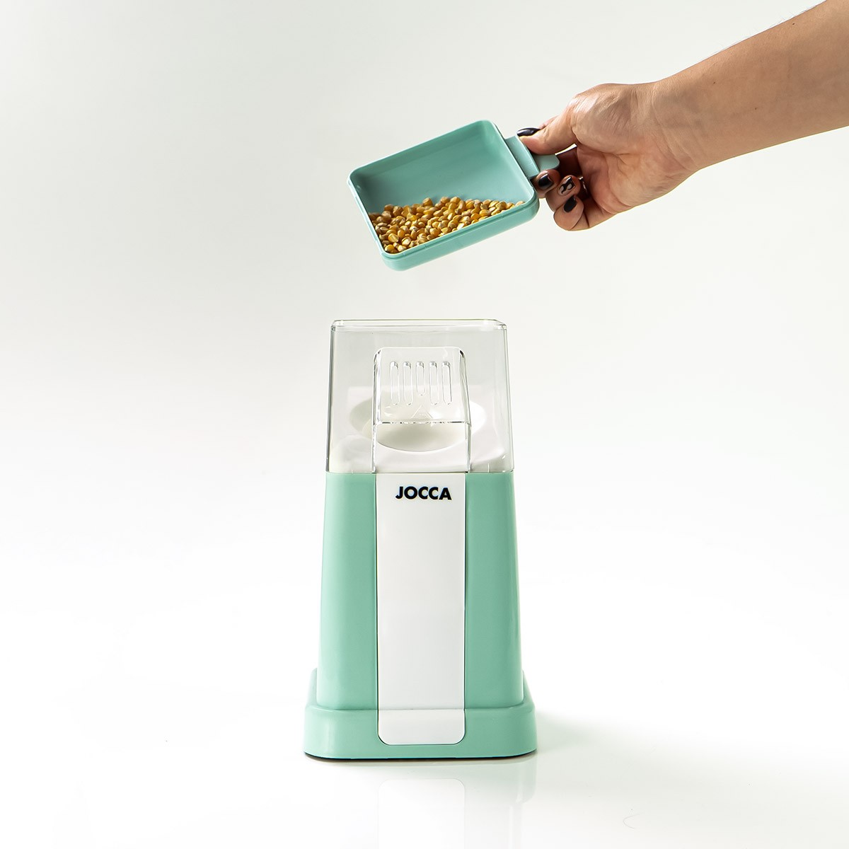 Una mano versa chicchi di mais da un misurino verde acqua in una macchina per popcorn verde acqua e bianca. Lo sfondo è bianco.