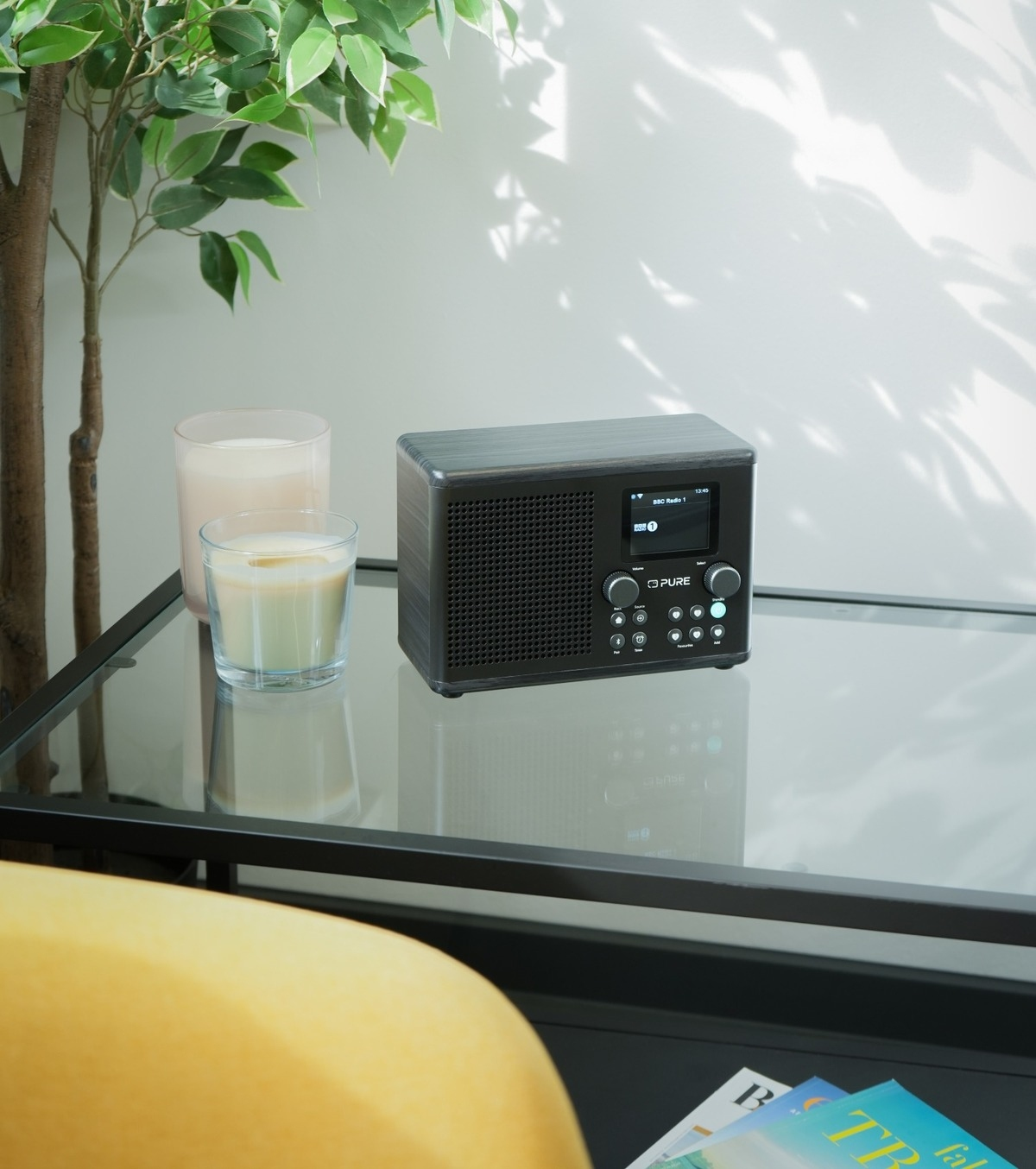 Ein schwarzes Radio auf einem Glastisch. Daneben zwei Kerzen in Glashaltern. Grüne Pflanze und gelber Stuhl im Hintergrund.