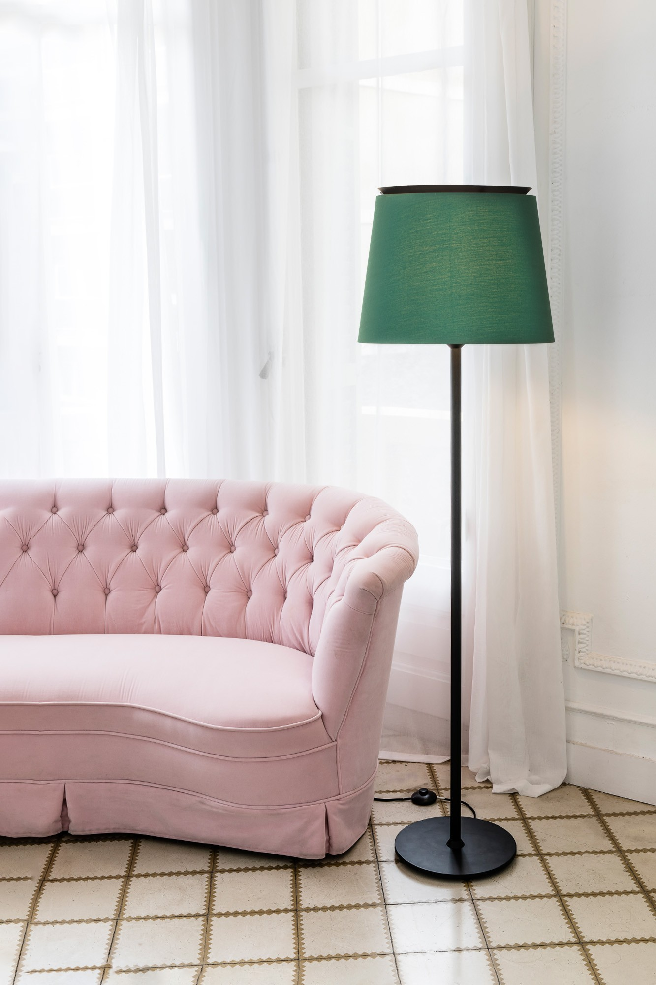 Un divano rosa trapuntato è accanto a una lampada nera con un paralume verde. Tende bianche e pavimento piastrellato sullo sfondo.