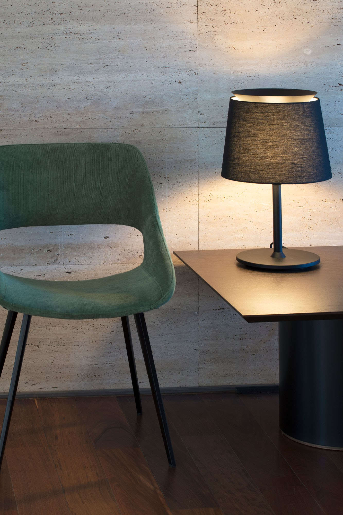Una sedia verde, lampada nera su tavolo e muro strutturato nell'immagine. Pavimento in legno in primo piano.