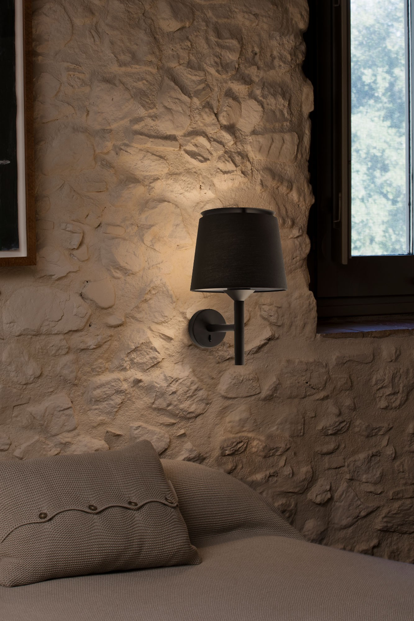 Una lampada da parete nera è montata su un muro di pietra. La lampada illumina un letto con cuscini. Una finestra è sullo sfondo.