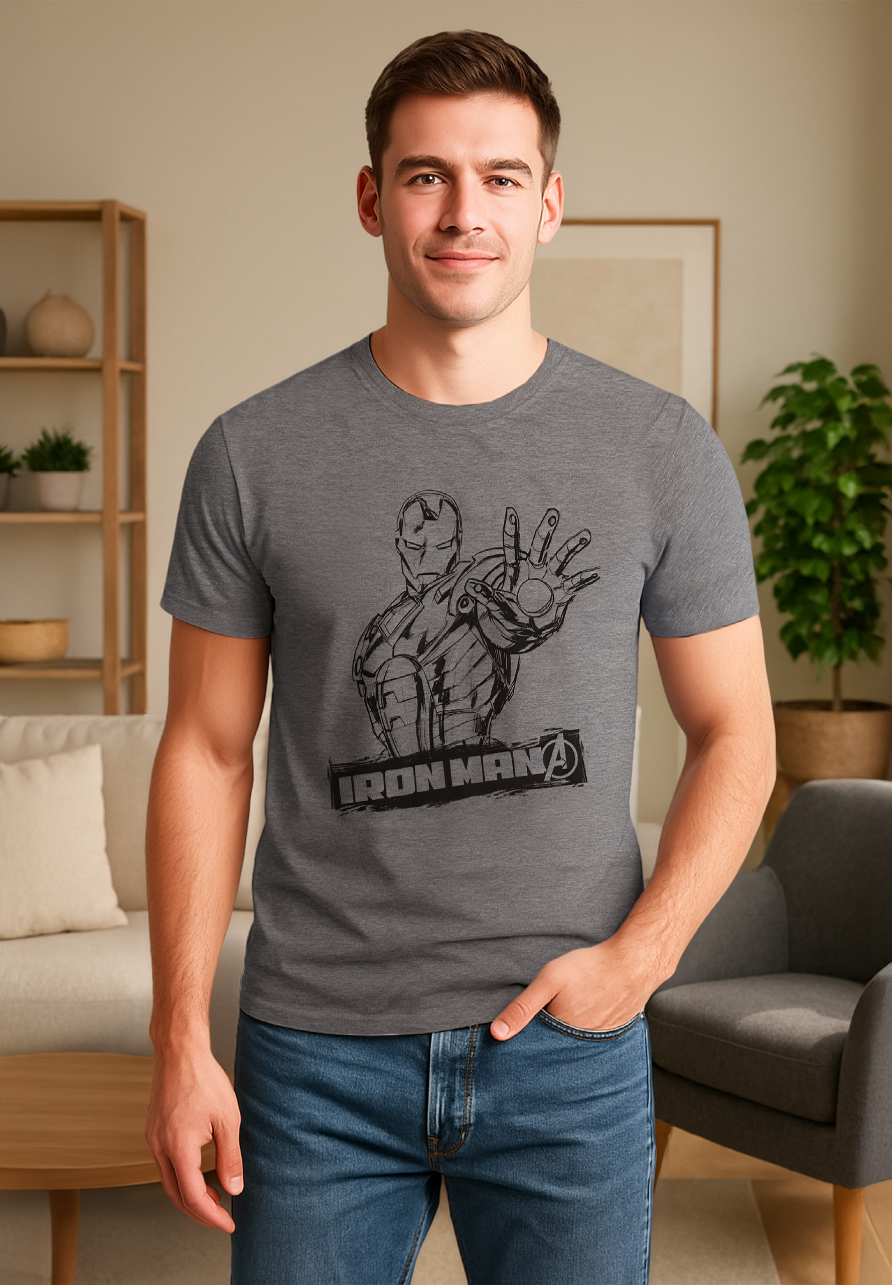 Ein Mann in einem grauen Iron-Man-Shirt und Jeans lächelt und steht in einem Wohnzimmer. Der Hintergrund umfasst Möbel und eine Pflanze.