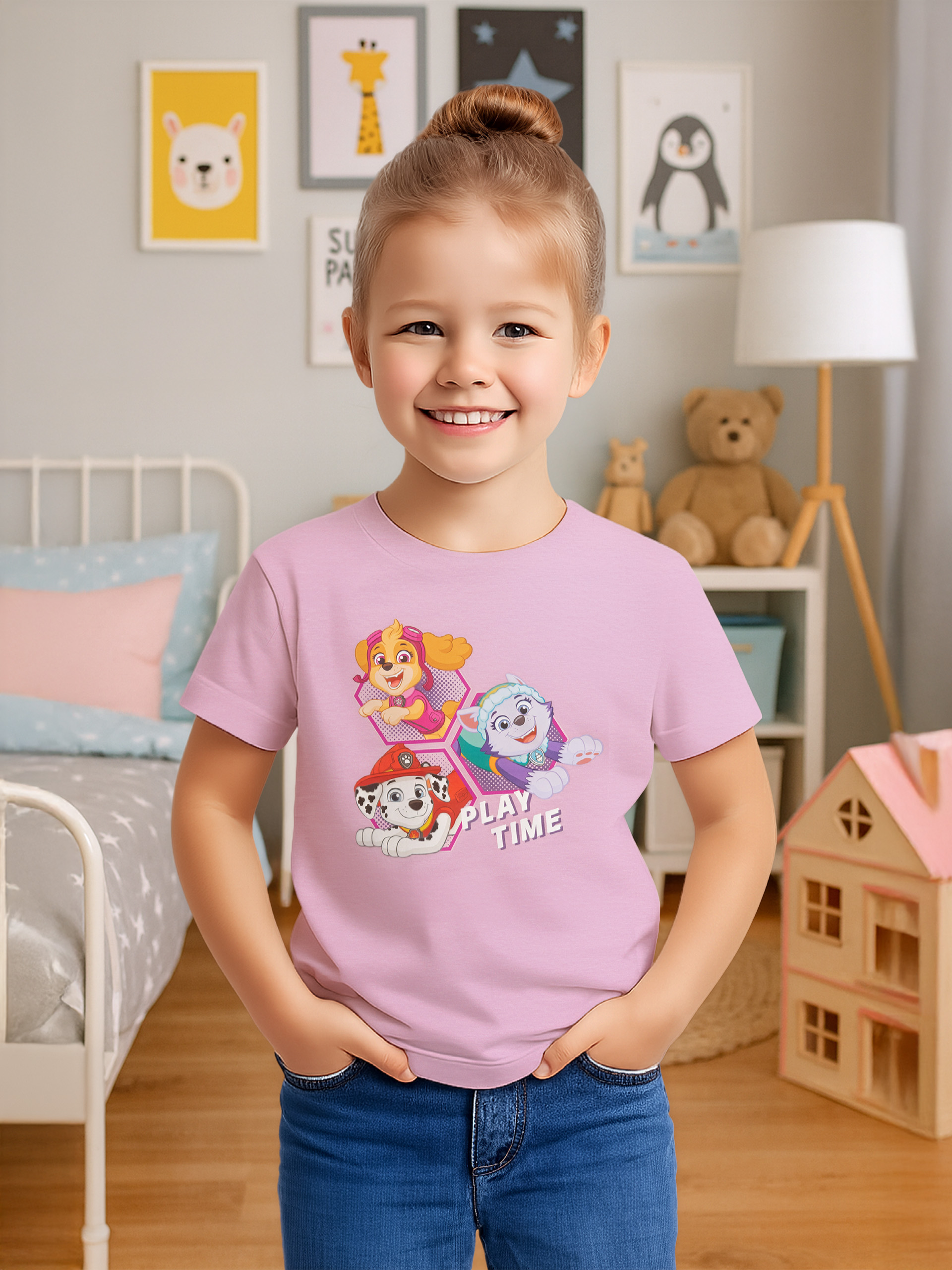 Ein Mädchen in einem rosa Shirt mit Paw Patrol-Charakteren lächelt. Sie trägt Jeans und steht in einem Zimmer.