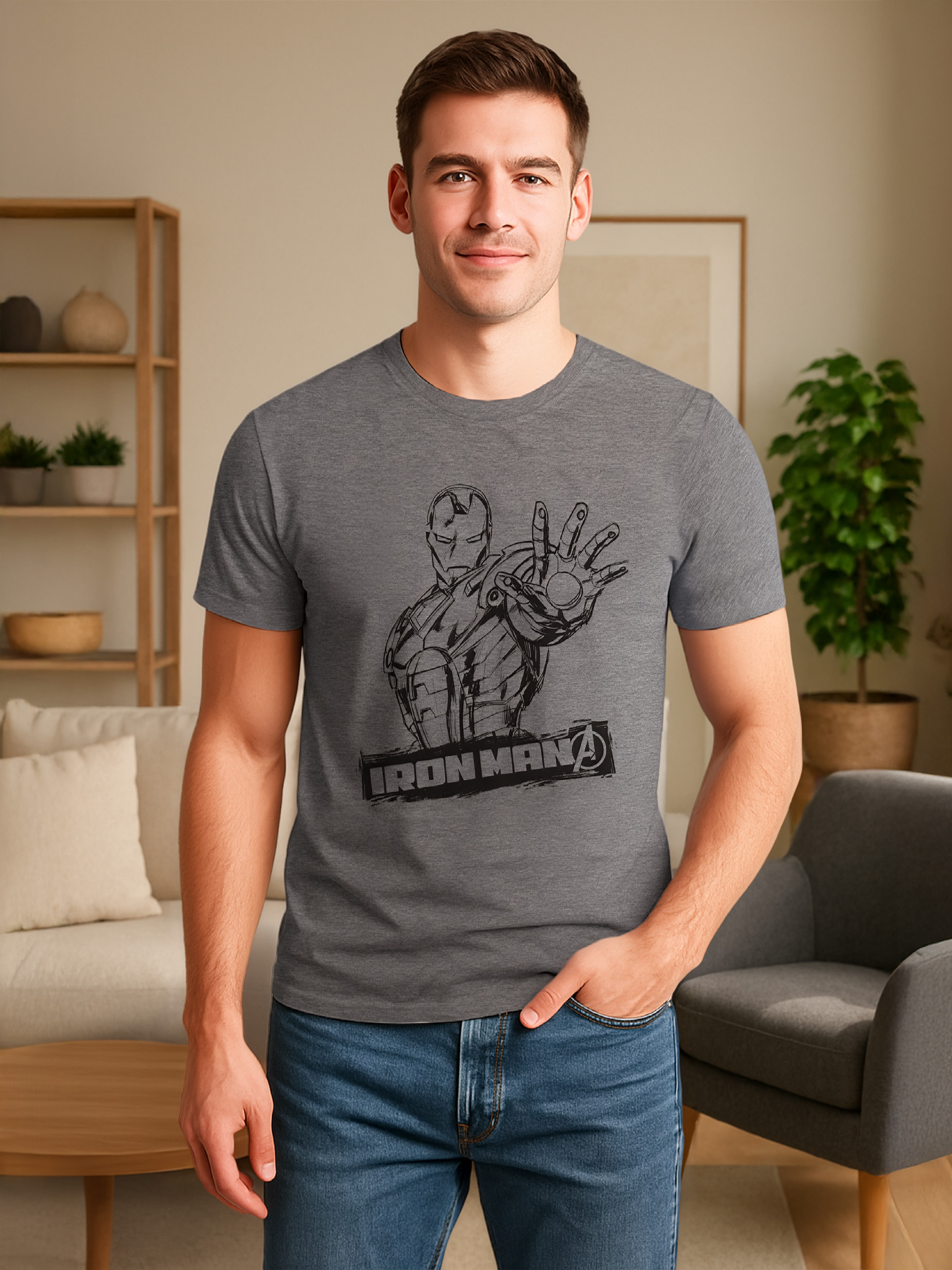Mann trägt graues Iron-Man-Shirt, Jeans, lächelt. Hintergrund: Wohnzimmer mit Sofa, Stuhl, Pflanzen.