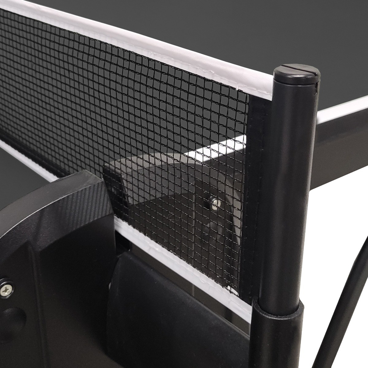 Close-up of a ping pong net. The net is black, framed with white. A black pole supports the net. Parts of the table are visible.