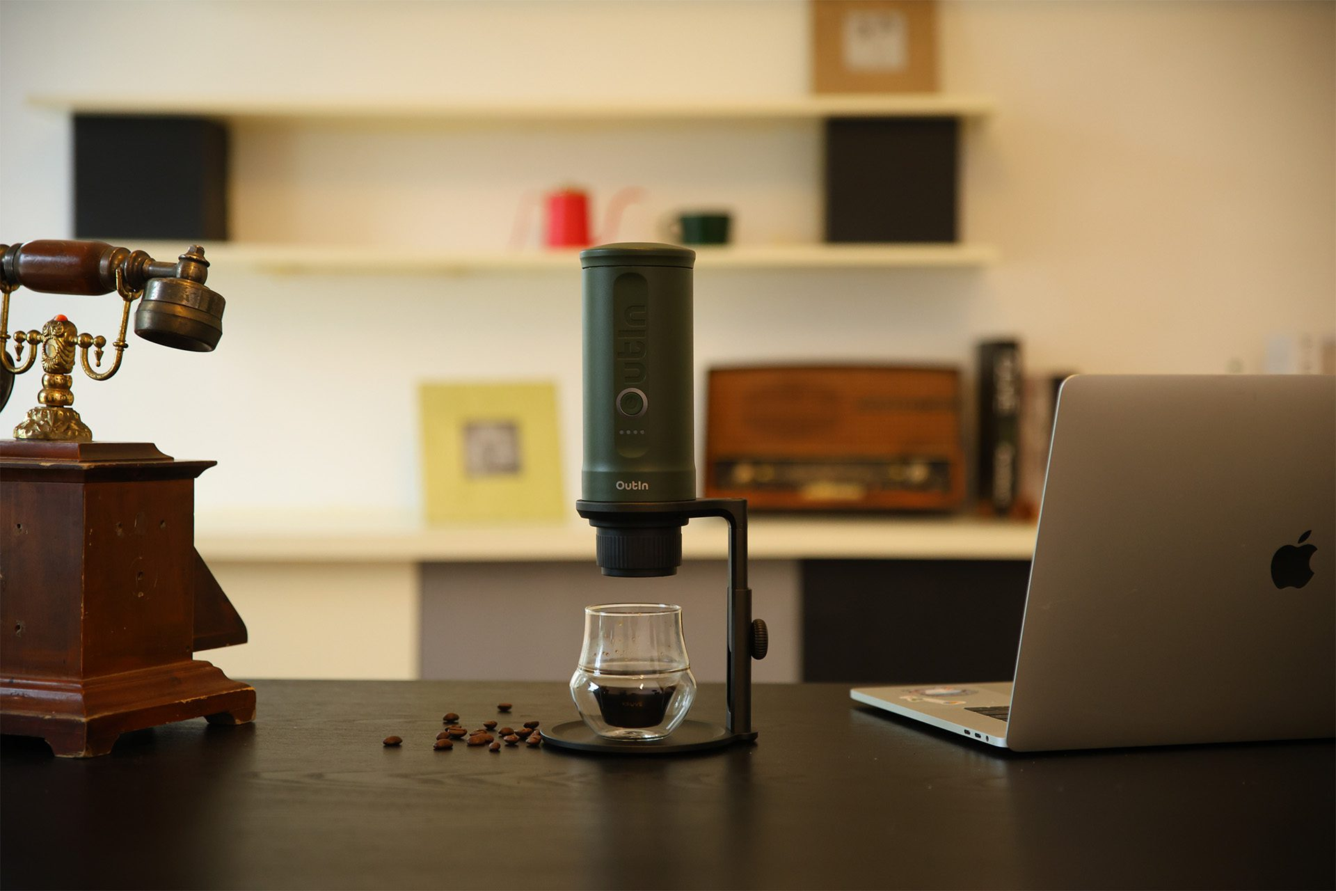 Ekspres do kawy ze szklanką na stole. Obok stary telefon i laptop. Ziarna kawy są rozrzucone.