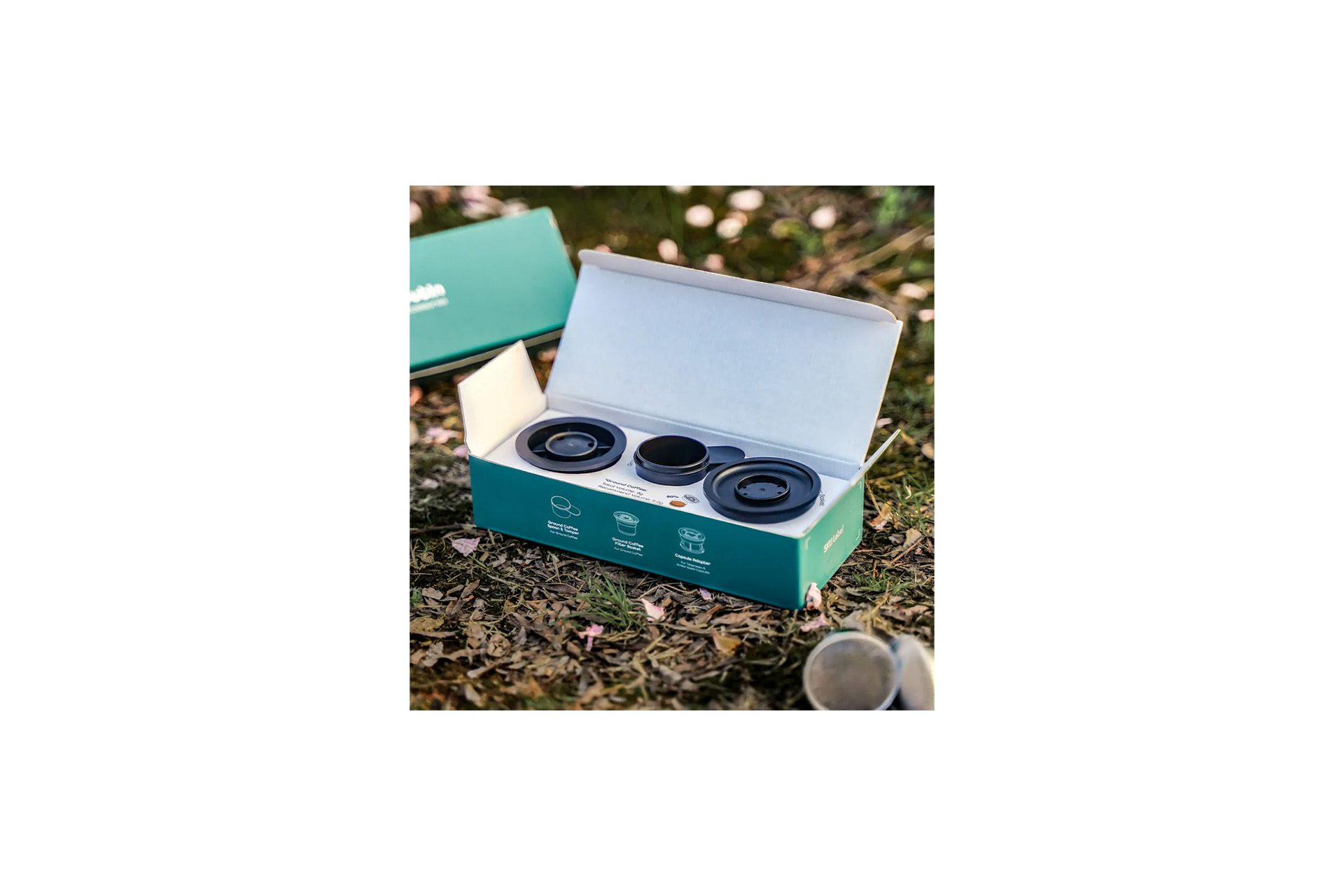 Geöffnete Schachtel mit schwarzen Kaffeepads, grüne Schachtel und Hintergrund aus Blättern und Blütenblättern.