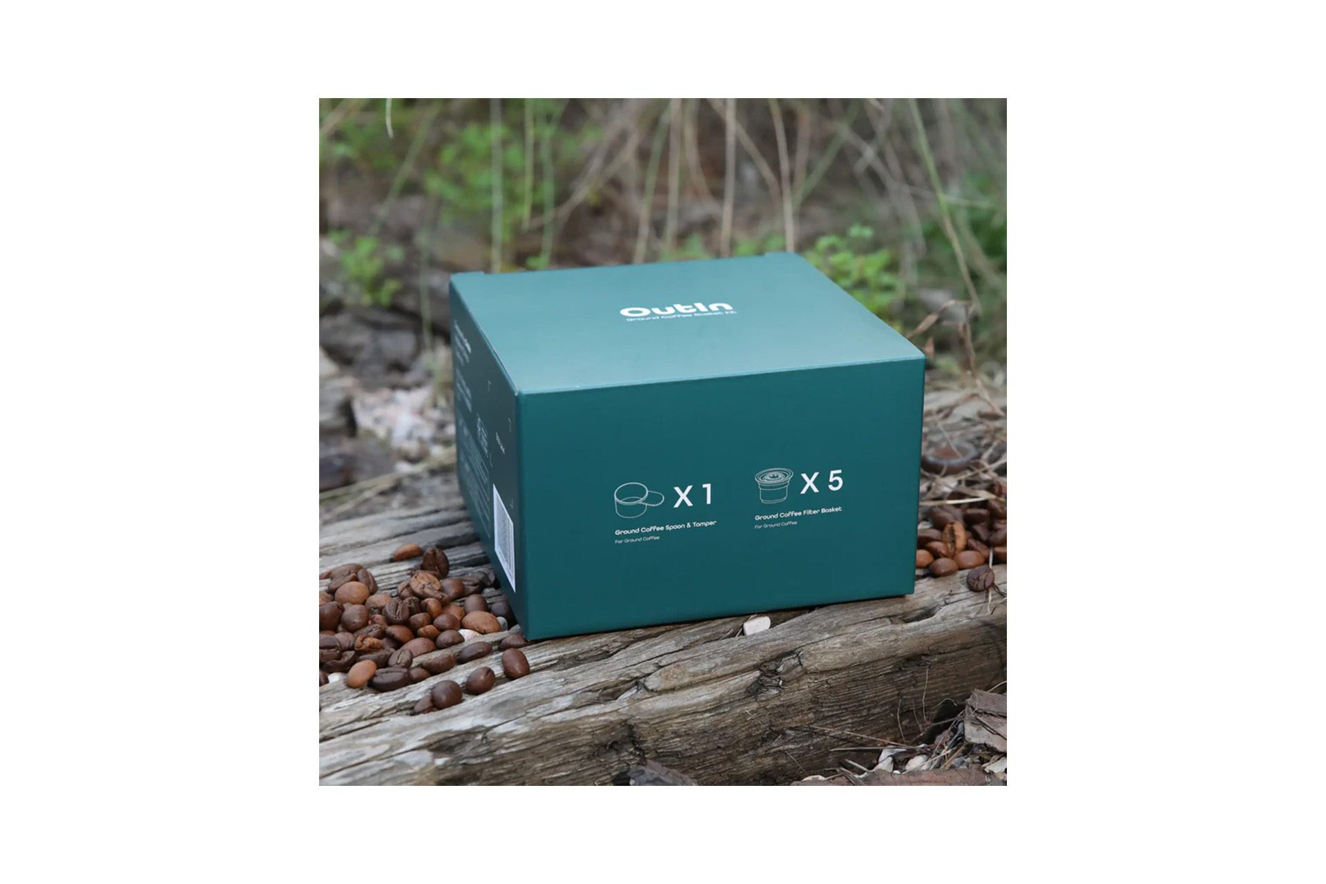 Eine teal Outin-Box mit Kaffeezubehör steht auf Holz, umgeben von Kaffeebohnen. Der Hintergrund ist unscharfes Grün.
