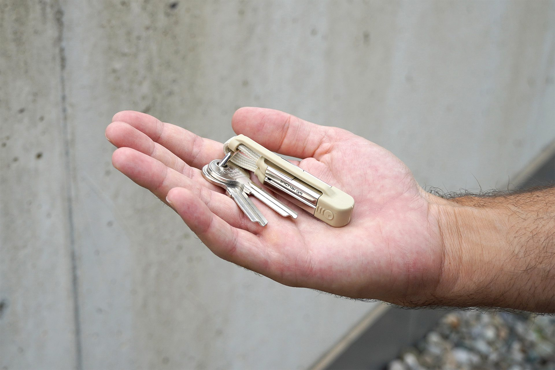 Een hand houdt twee sleutels en een 'inCharge'-apparaat vast. De achtergrond is een betonnen muur. Het apparaat en de sleutels zijn zilver en beige.