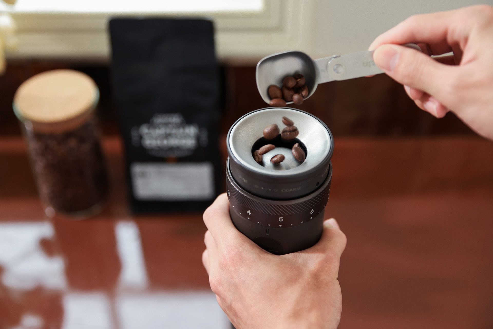 Osoba wsypuje ziarna kawy z łyżki do młynka do kawy. Słoik i torba kawy są w tle.