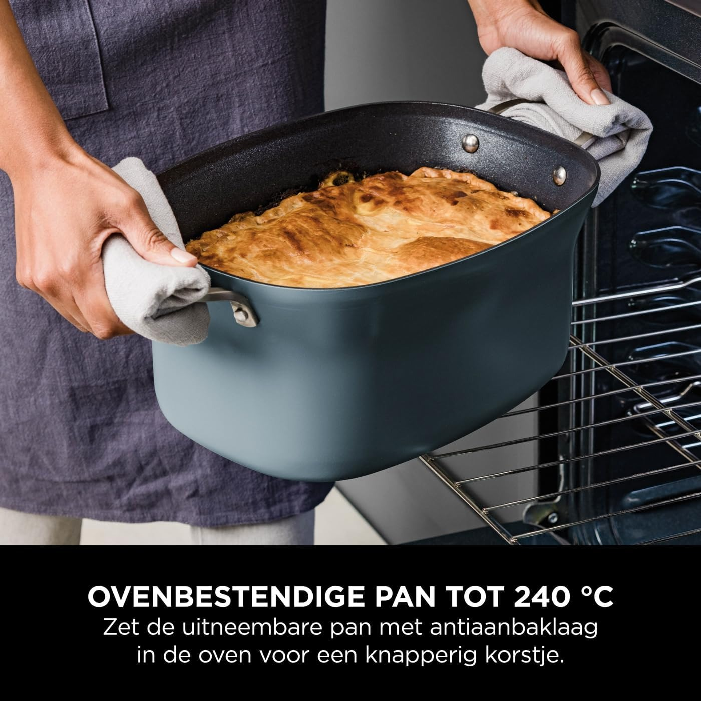 Handen houden een donkerblauwe bakvorm met een goudbruine taart vast. De pan wordt in de oven geplaatst.