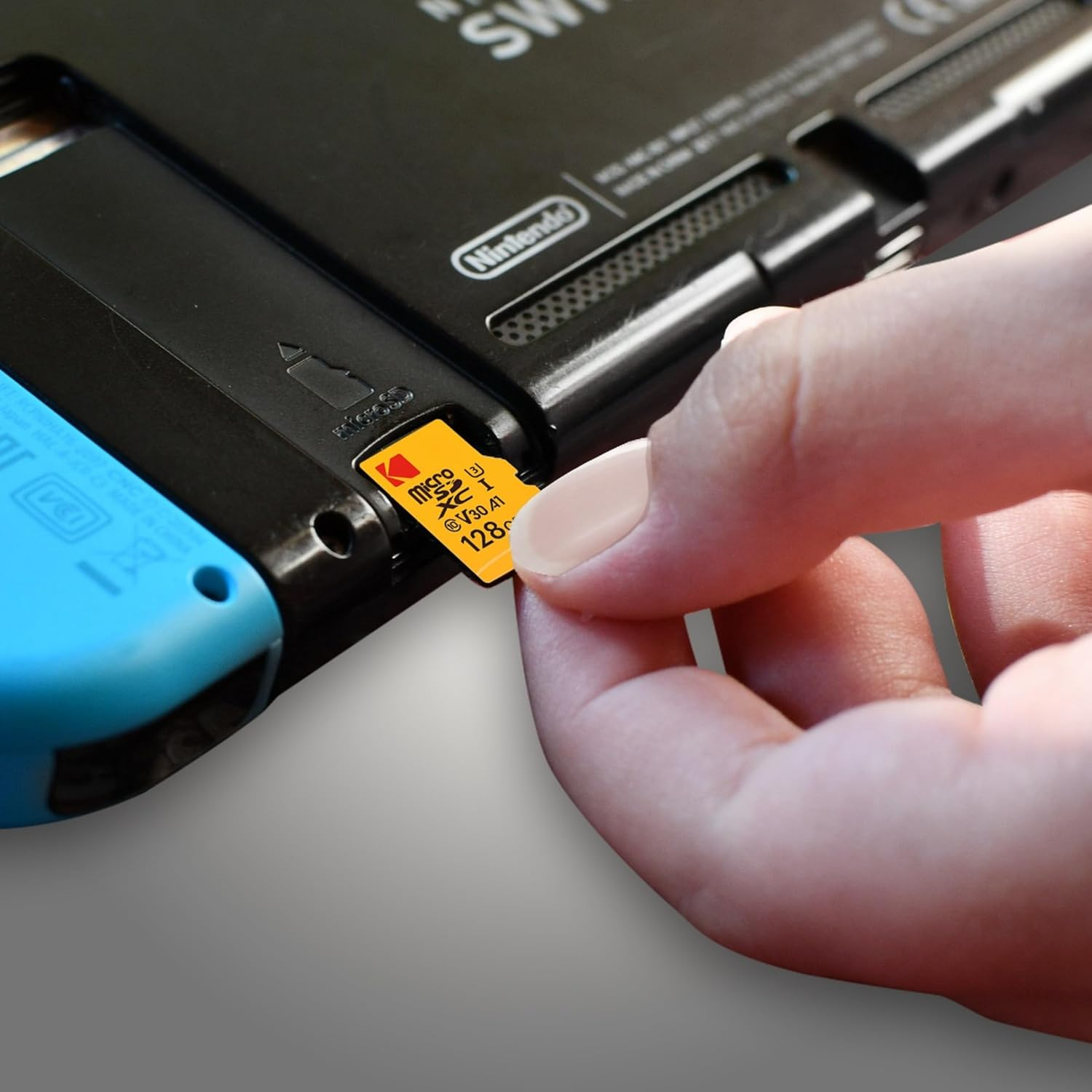 Eine Hand, die eine gelbe Kodak Micro-SD-Karte in eine schwarze Nintendo Switch einlegt. Die Konsole liegt auf einer grauen Oberfläche.