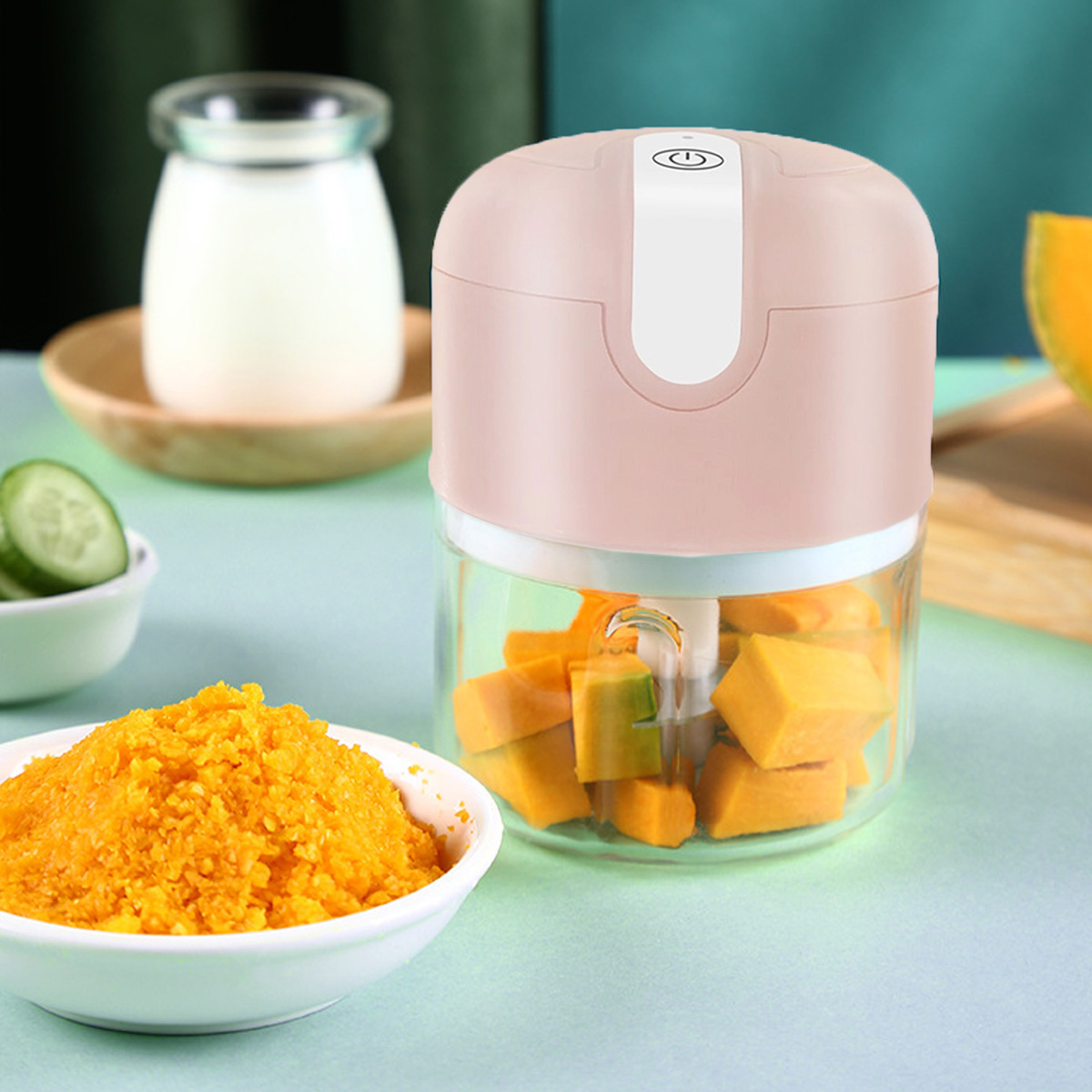 Ein Foodprozessor mit gehacktem Kürbis. Eine Schüssel mit orangefarbenem Püree und Gurkenscheiben. Ein Glas mit Flüssigkeit und ein Holzbrett im Hintergrund.